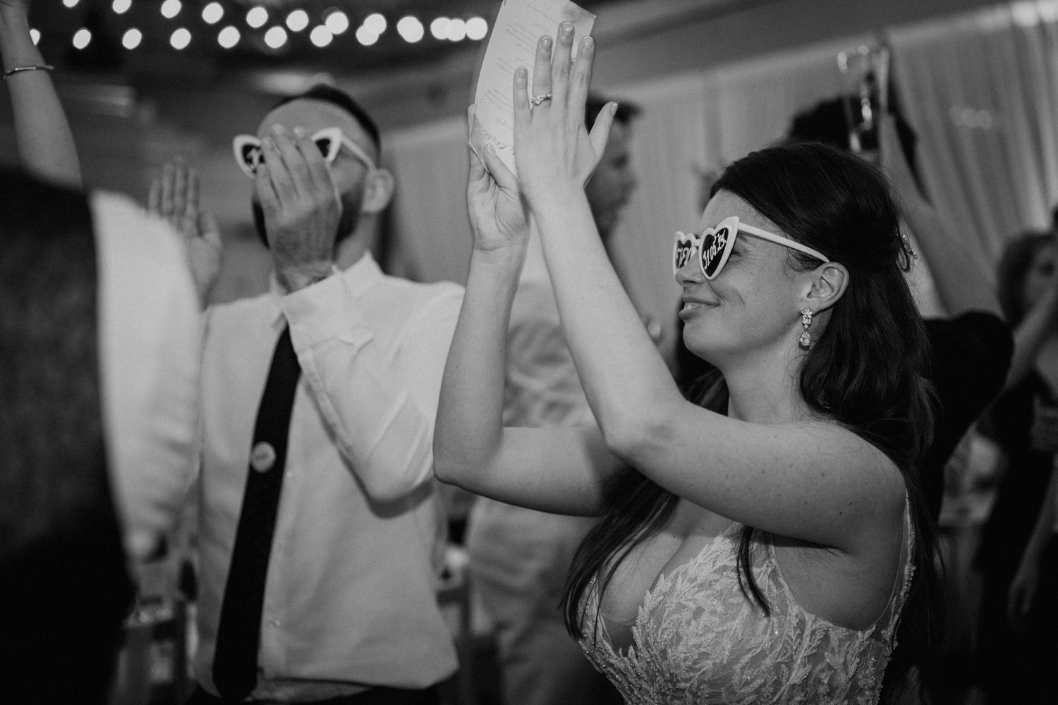 Black and white photo of wedding guests dancing and clapping hands wearing fun novelty glasses at a reception.