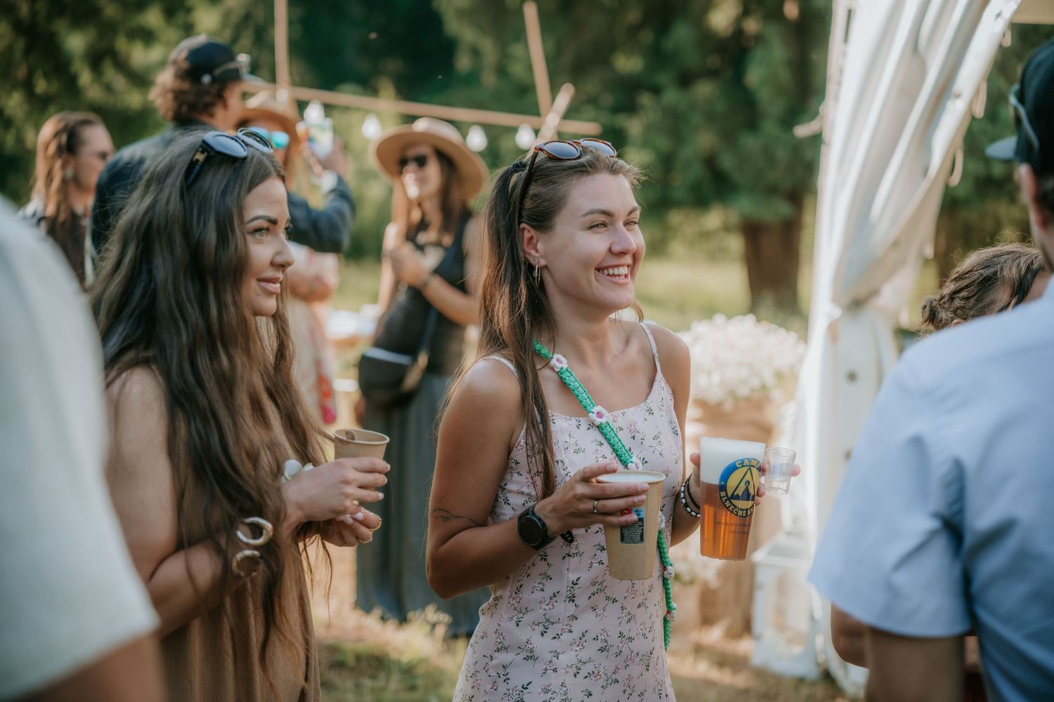 Two smiling women holding drinks at an outdoor summer festival, with friends gathered in the background.