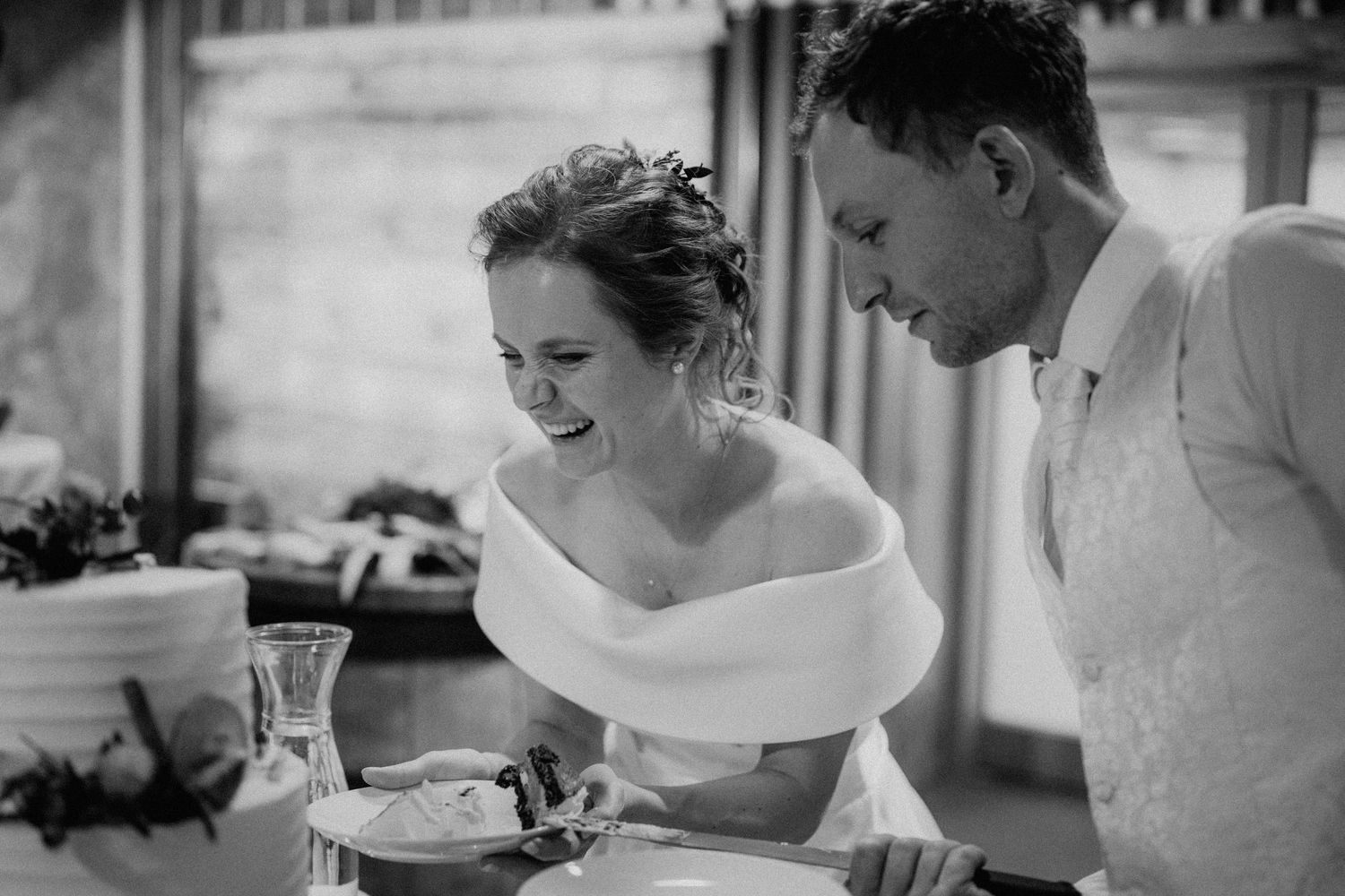 Black and white photo of a laughing bride in off-shoulder dress cutting wedding cake with groom.