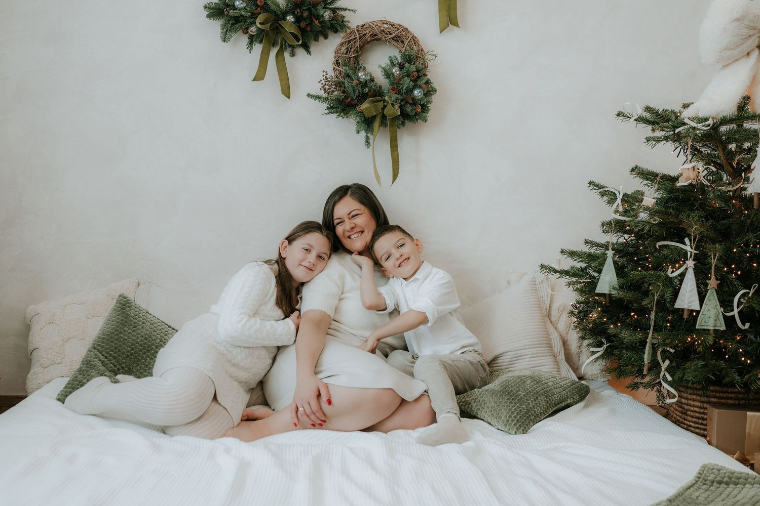 A mother in white sits on a bed hugging her two children near a Christmas tree and holiday decorations.