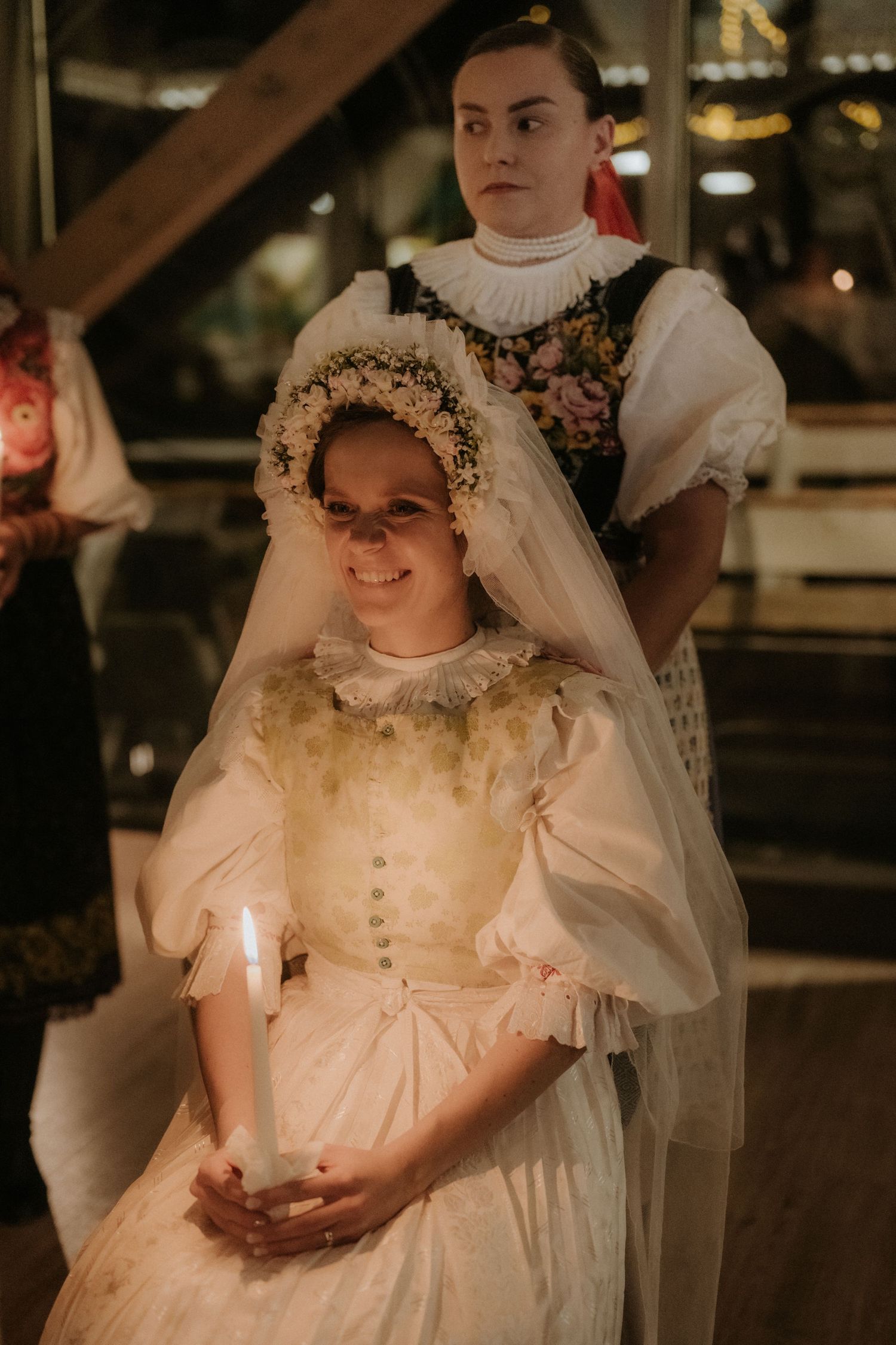 Young girl in vintage bridal gown holding candle with floral crown, standing before woman in traditional folk costume.