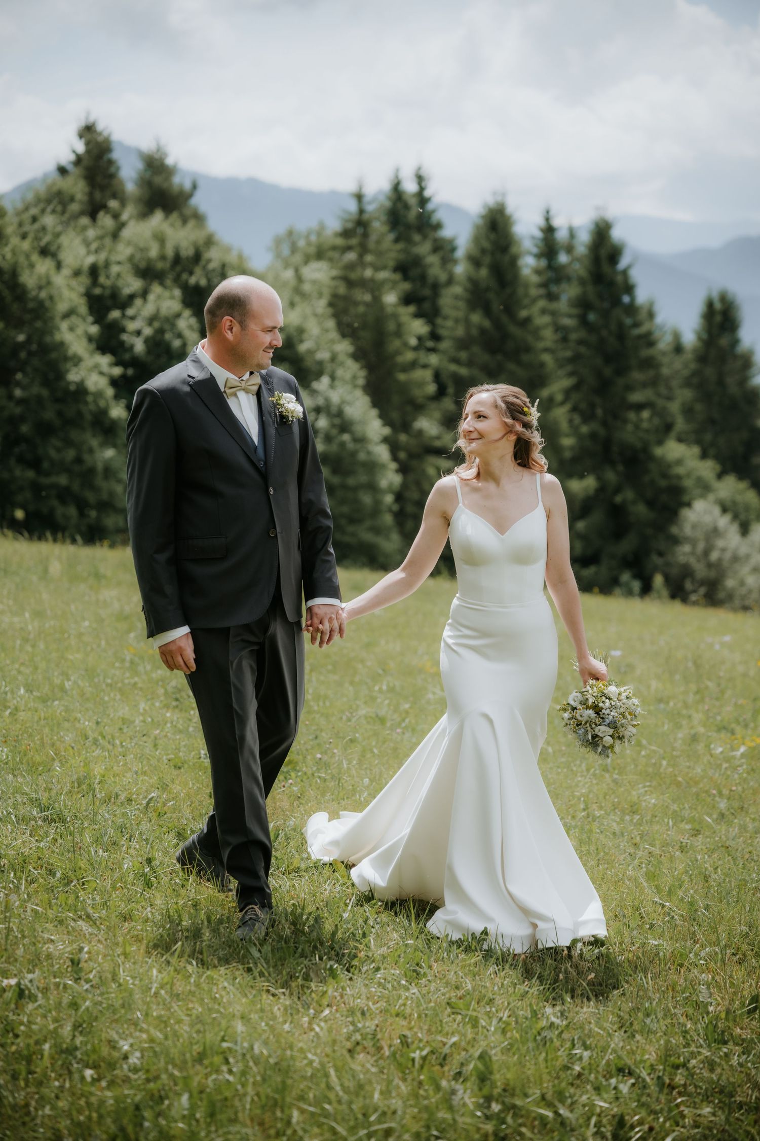 A bride and groom walk hand-in-hand through a lush green meadow with pine trees and mountains in the background.