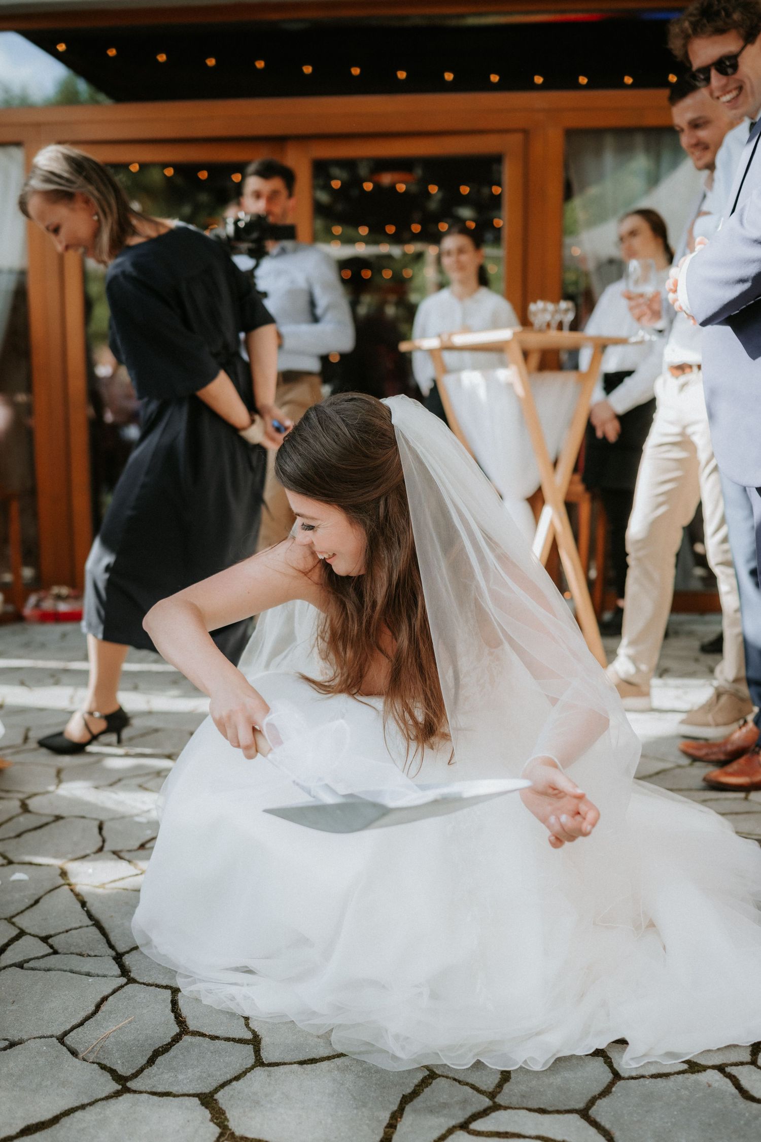 Bride in white gown and veil bending down to pick up an item on stone patio at outdoor wedding reception.
