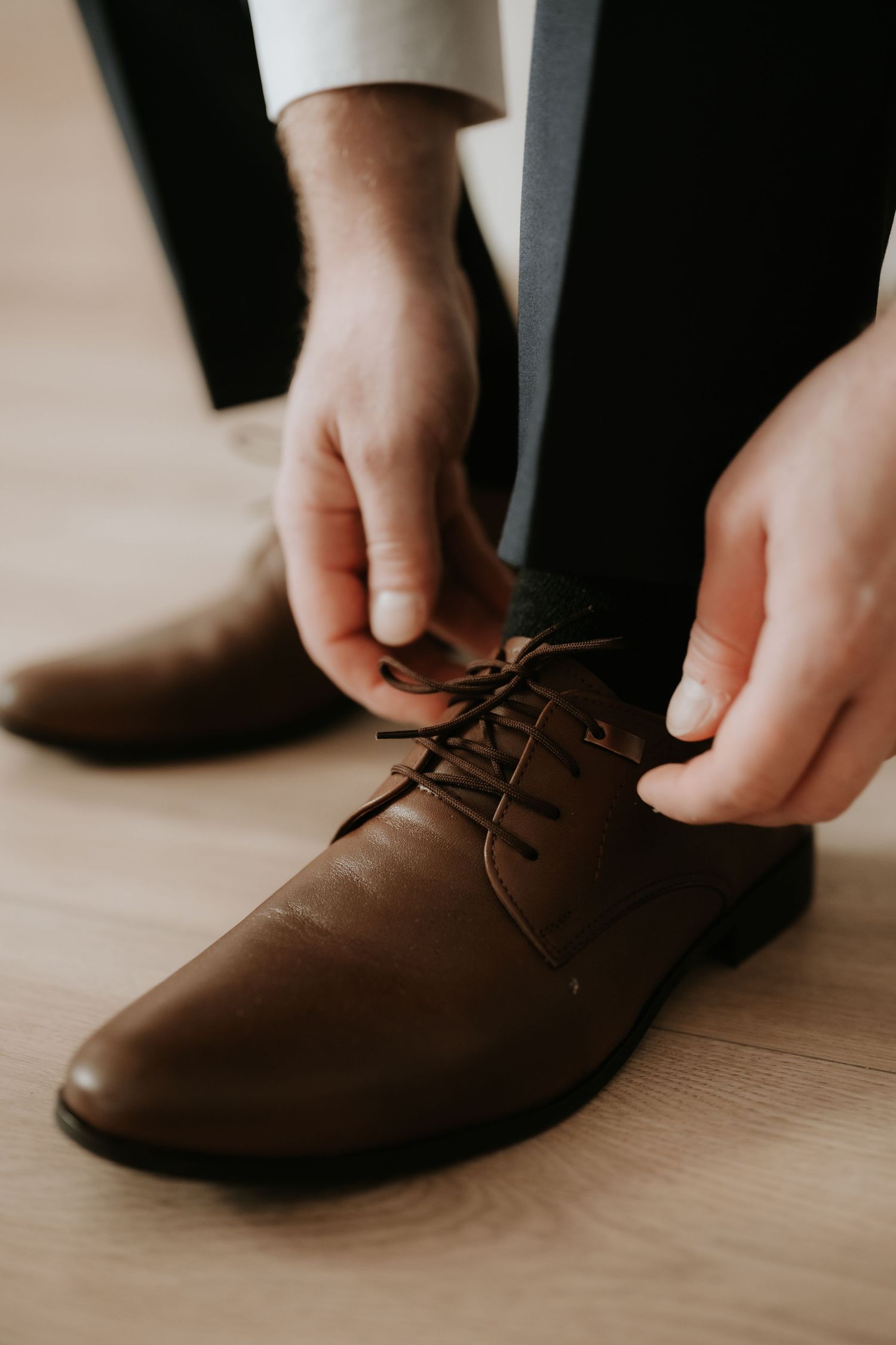 Man tying brown leather dress shoes with dark laces while wearing formal trousers on a wooden floor.
