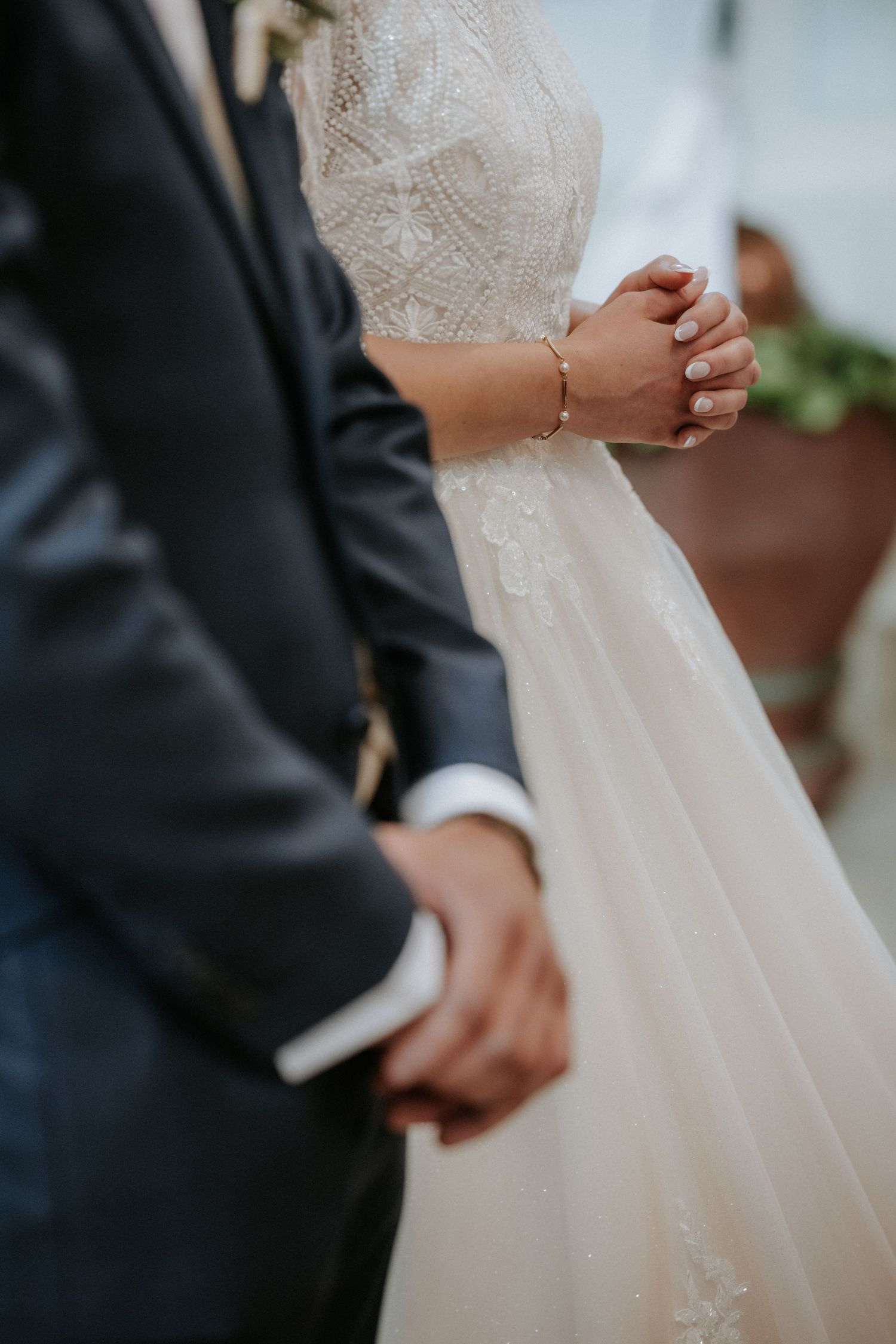 Bride and groom holding hands at wedding ceremony, bride in lace gown with bracelet, groom in dark suit.