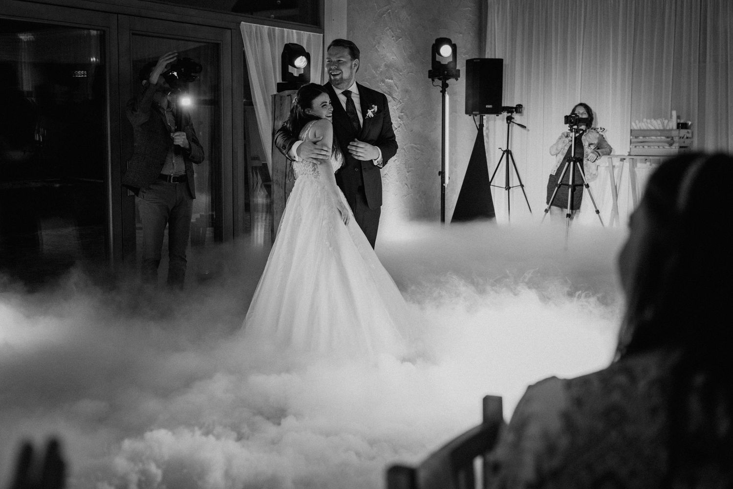 Black and white photo of bride and groom sharing first dance surrounded by dramatic fog at elegant wedding reception.