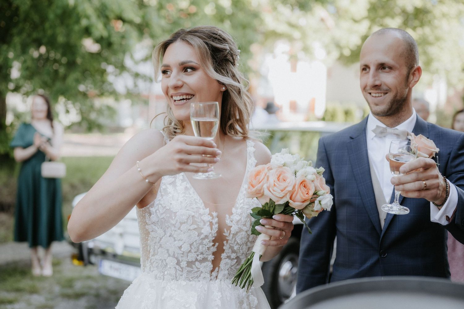 Bride and groom toasting champagne outdoors, bride holding pink rose bouquet in elegant lace wedding dress.