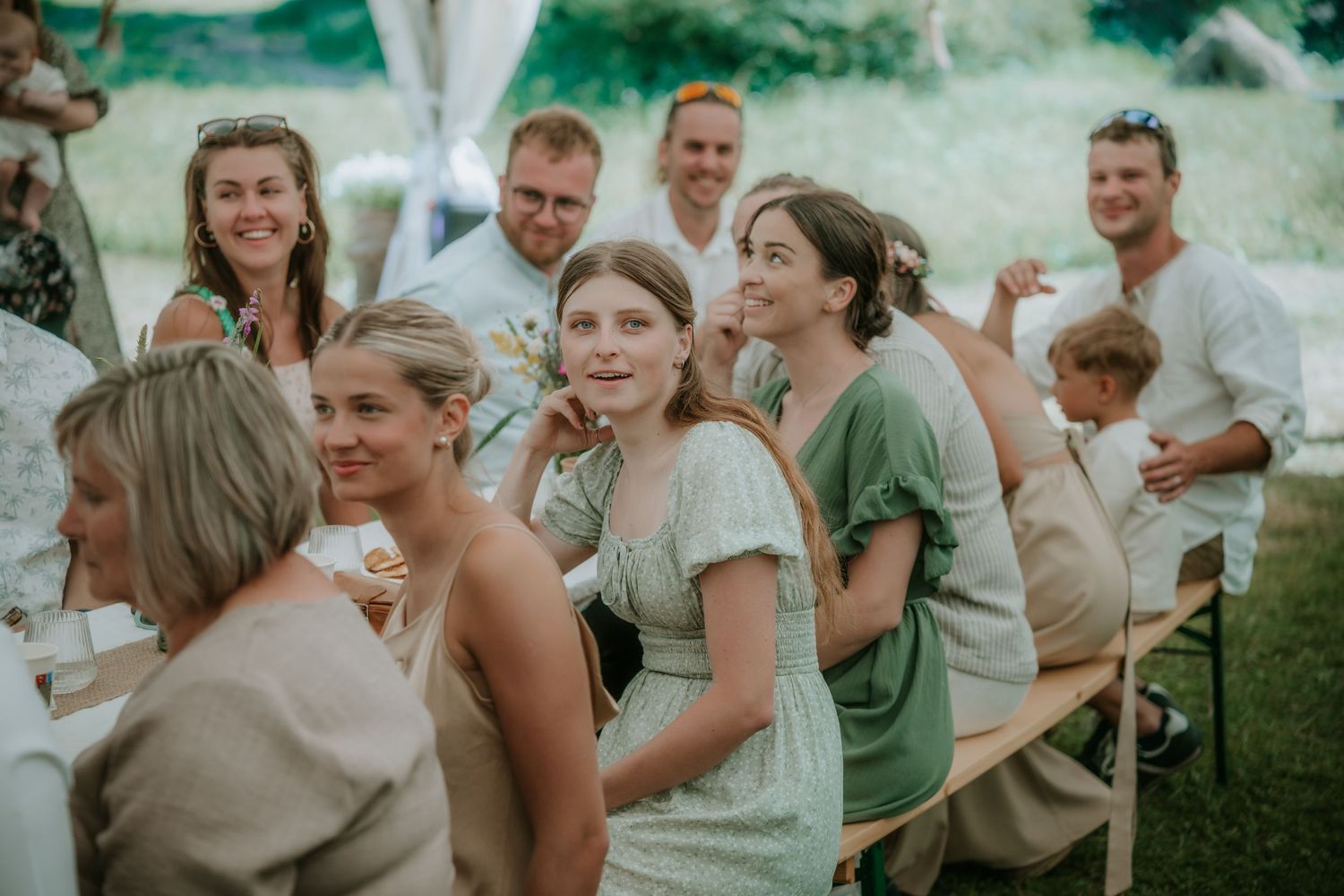 Guests laughing and enjoying an outdoor wedding reception seated at long wooden tables under a white tent.