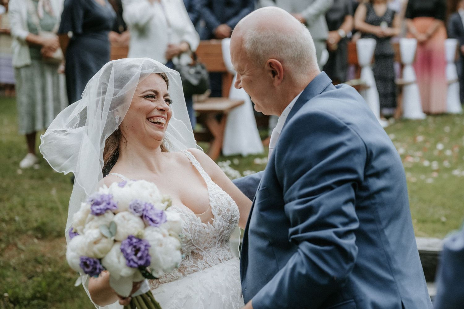 Smiling bride in lace gown and veil holds purple bouquet while exchanging vows with groom in blue suit outdoors.