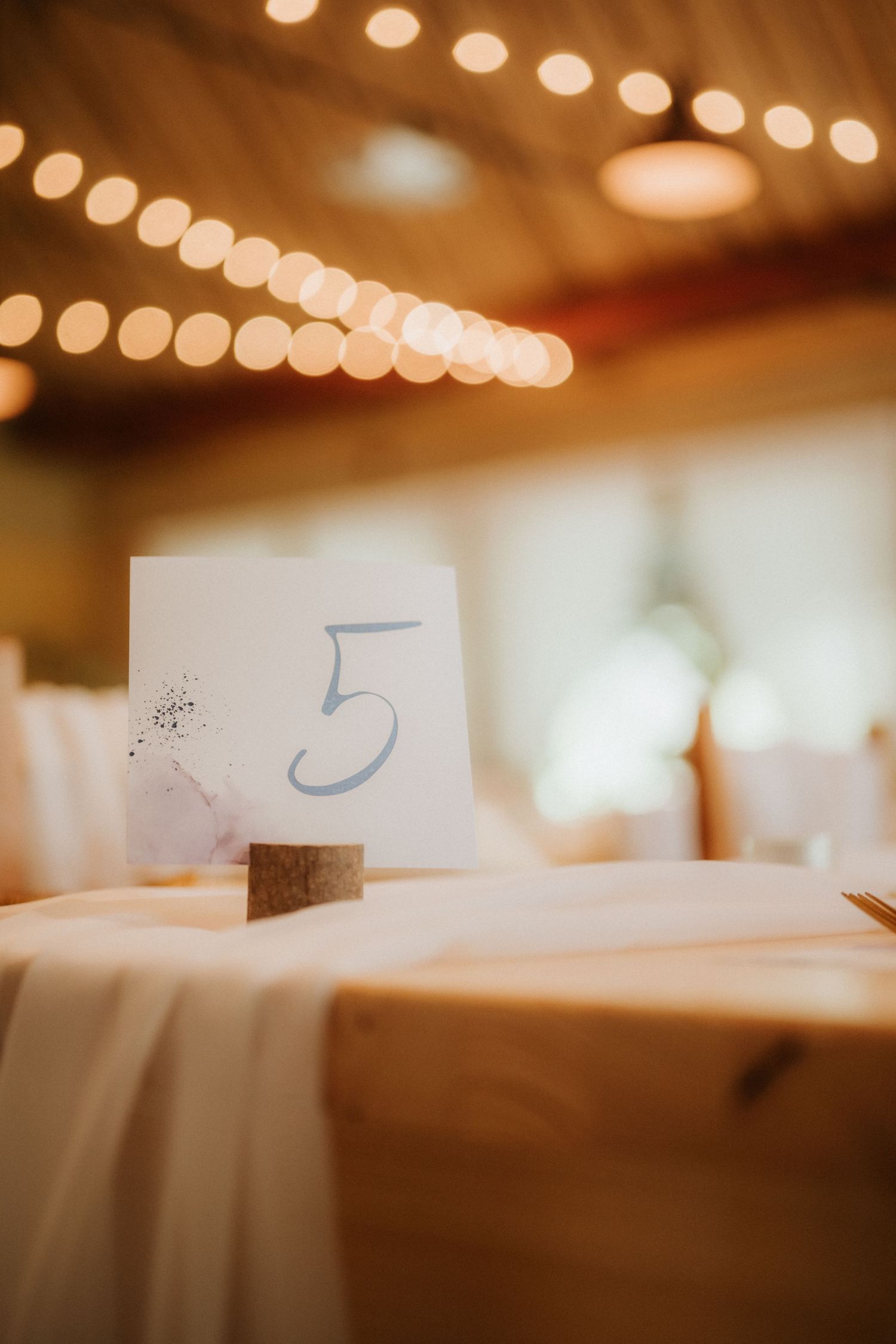 Table number five card in wooden holder at elegant wedding reception with warm string lights glowing in background.