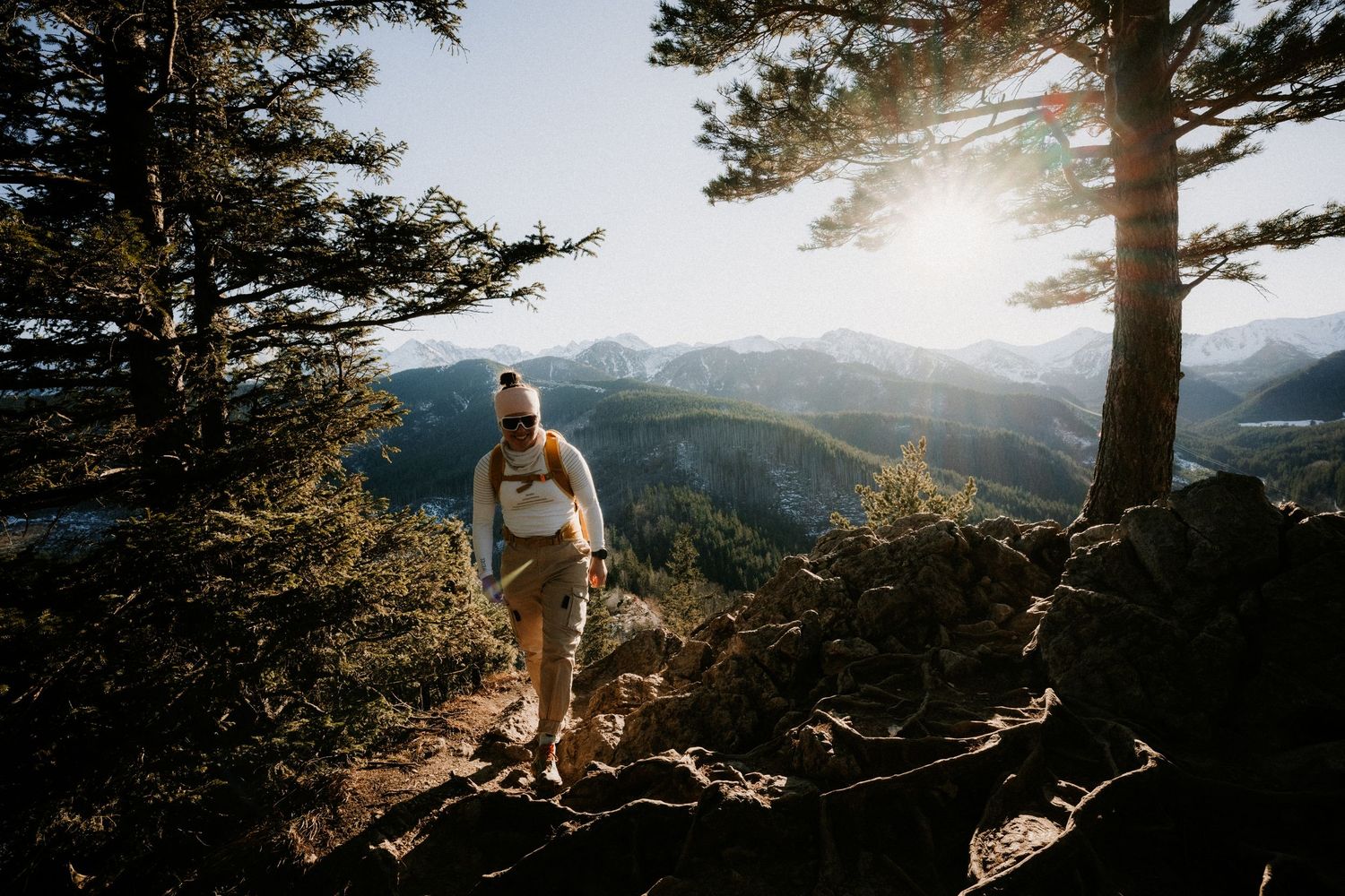 Hiker walking through pine trees on rocky mountain trail with stunning valley views at sunset.