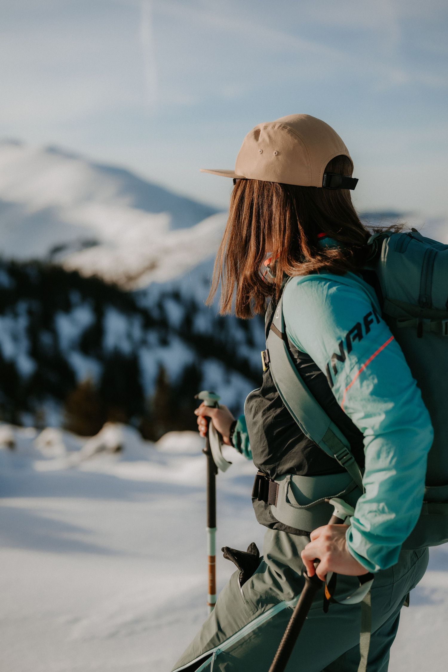 Female backcountry skier in teal jacket and tan cap gazing at snow-covered mountain terrain.