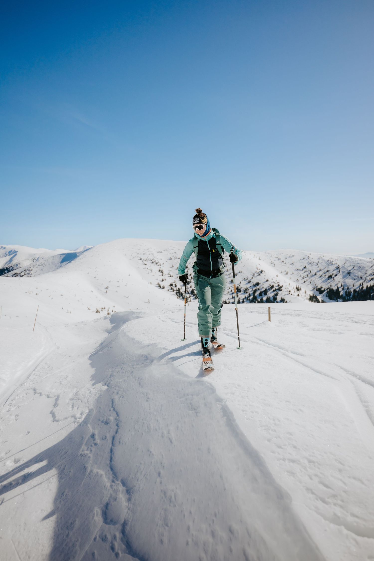 Female skier in teal ski suit smiling while ski touring up a snowy mountain on a sunny day.