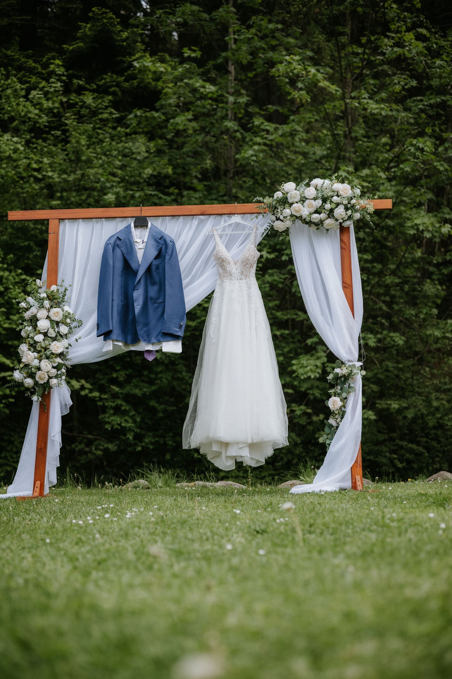 Wedding dress and blue suit jacket hanging on a floral arch outdoors with white draping and green trees.