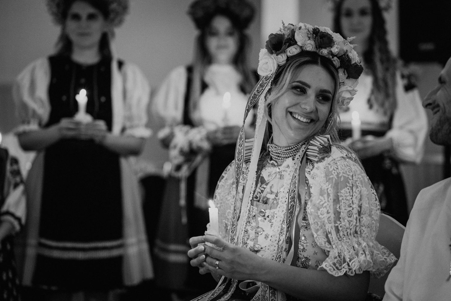 Black and white photo of a smiling woman in traditional folk costume with floral crown at a cultural ceremony.