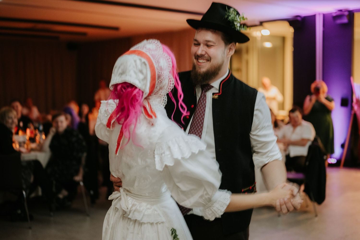 Couple dancing in traditional folk costumes at a wedding reception, man in black hat and vest, woman in white dress.
