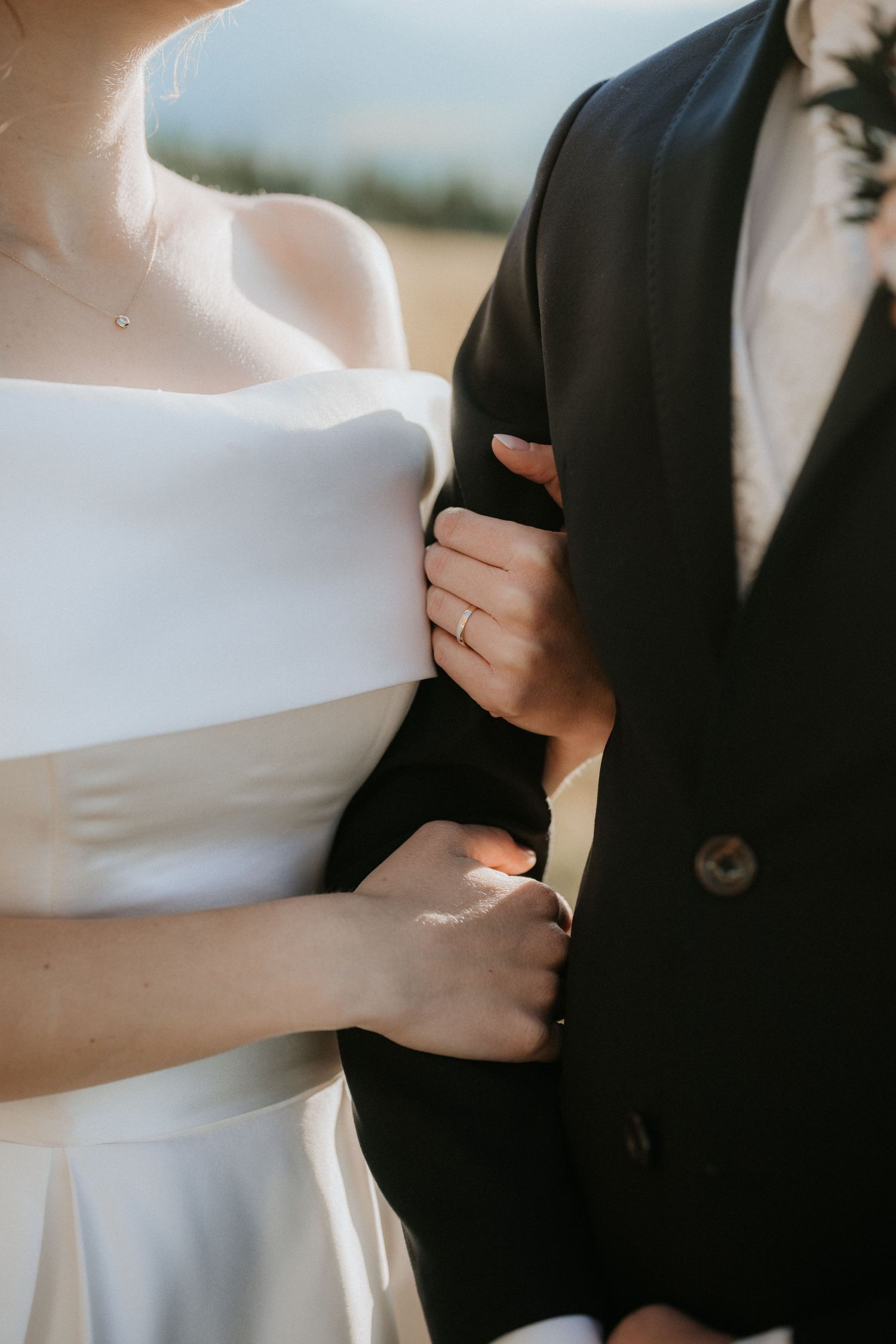 Close-up of bride in white off-shoulder dress holding grooms black suit jacket with wedding ring visible.