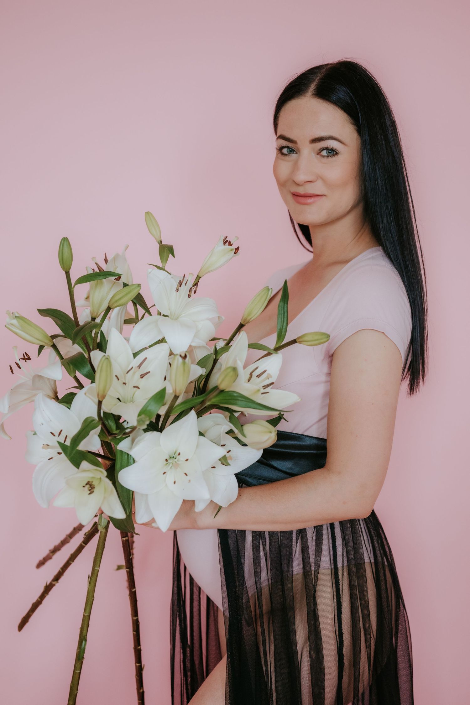 Smiling dark-haired woman in pink top and black tulle skirt holding white lilies against pink background.