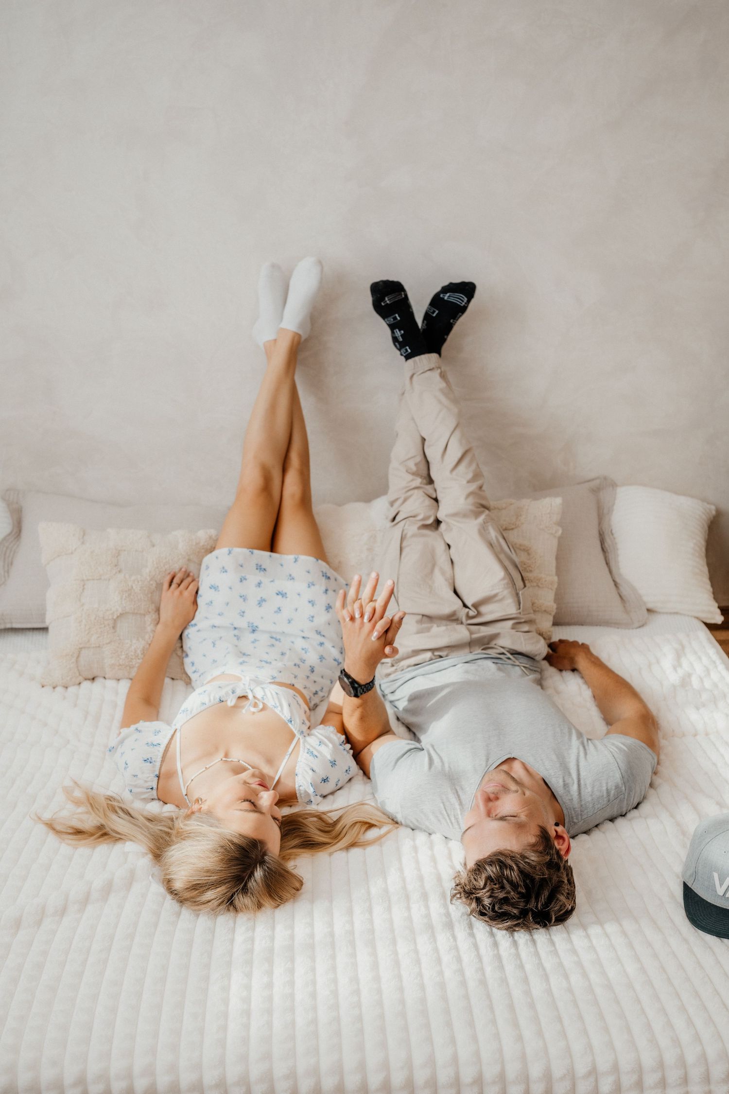 A happy couple lies on a white bed with their legs raised, laughing and holding hands in a bright cozy bedroom.