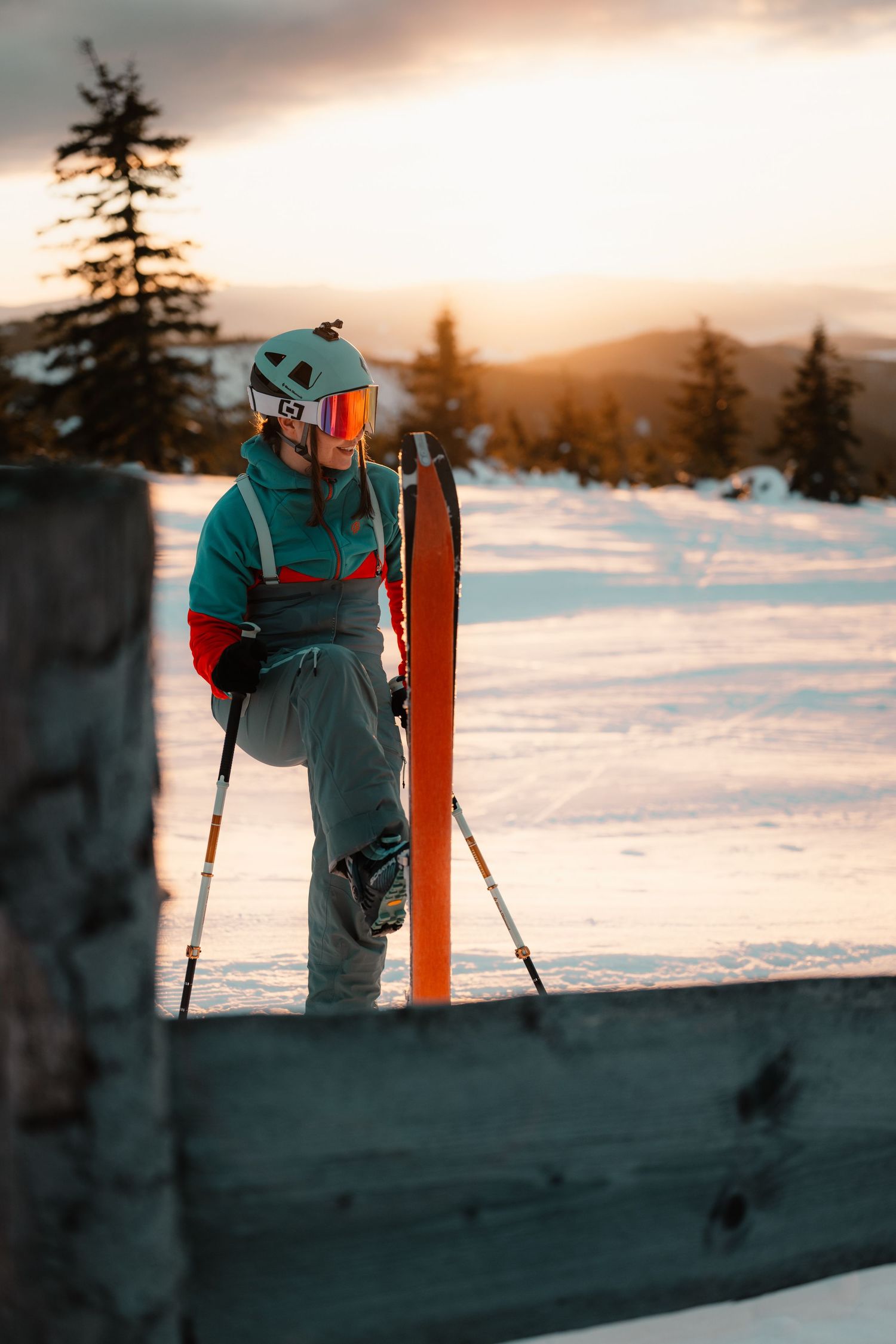 Female skier leaning on orange skis at snowy mountain resort during warm golden sunset.