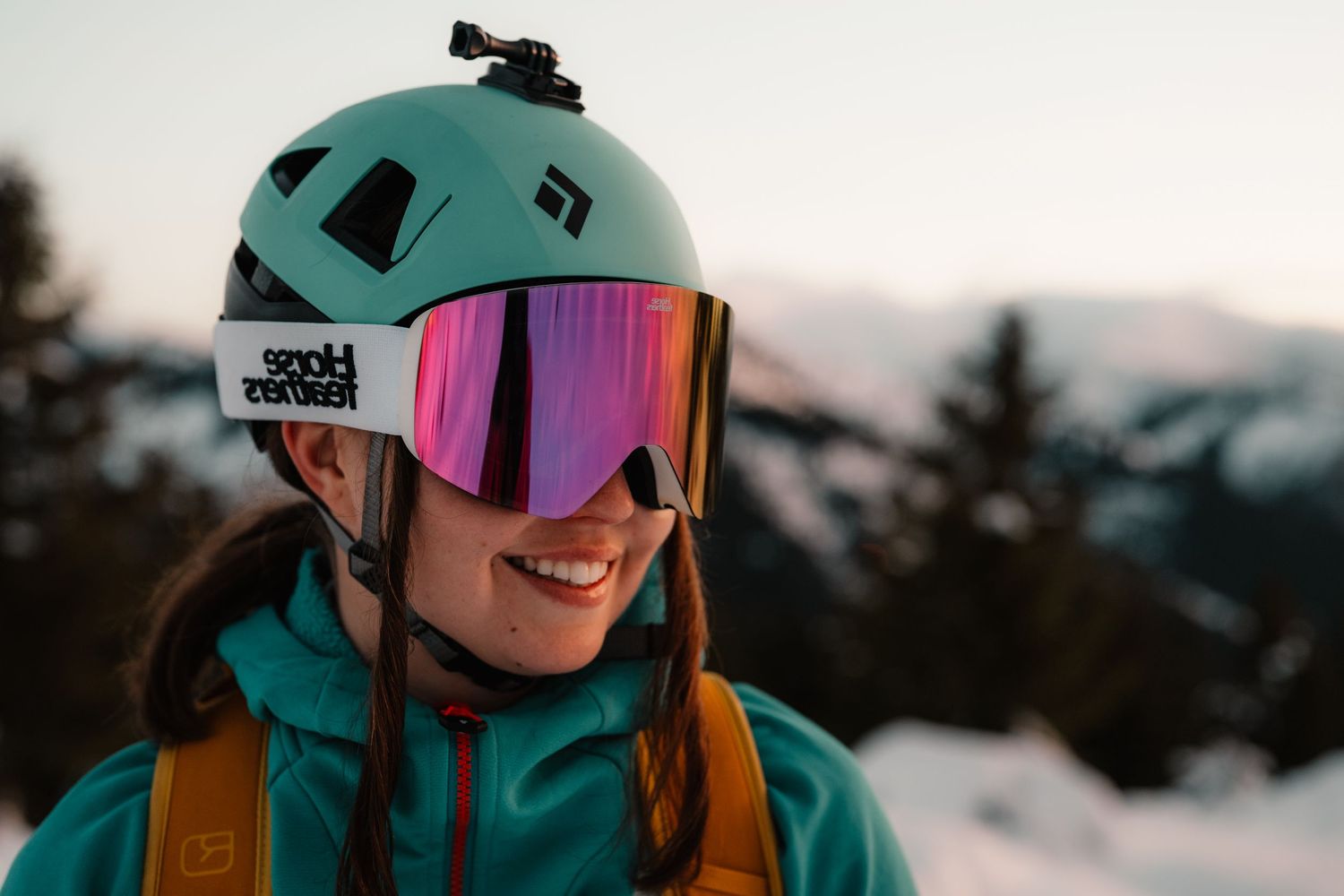 Close-up of smiling woman wearing teal Black Diamond helmet with pink mirrored ski goggles outdoors.