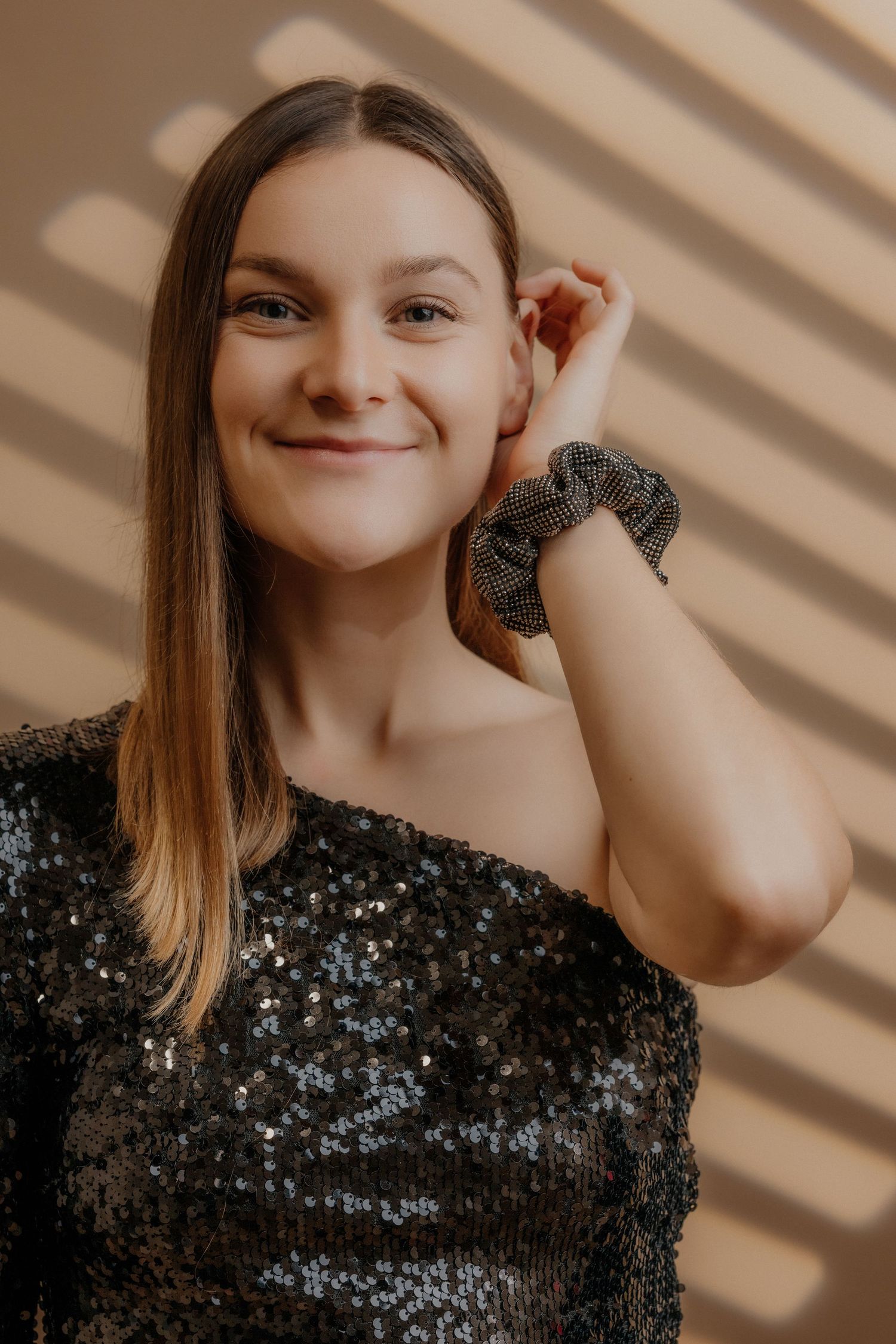 Smiling young woman in black sequin top touching hair with scrunchie on wrist.