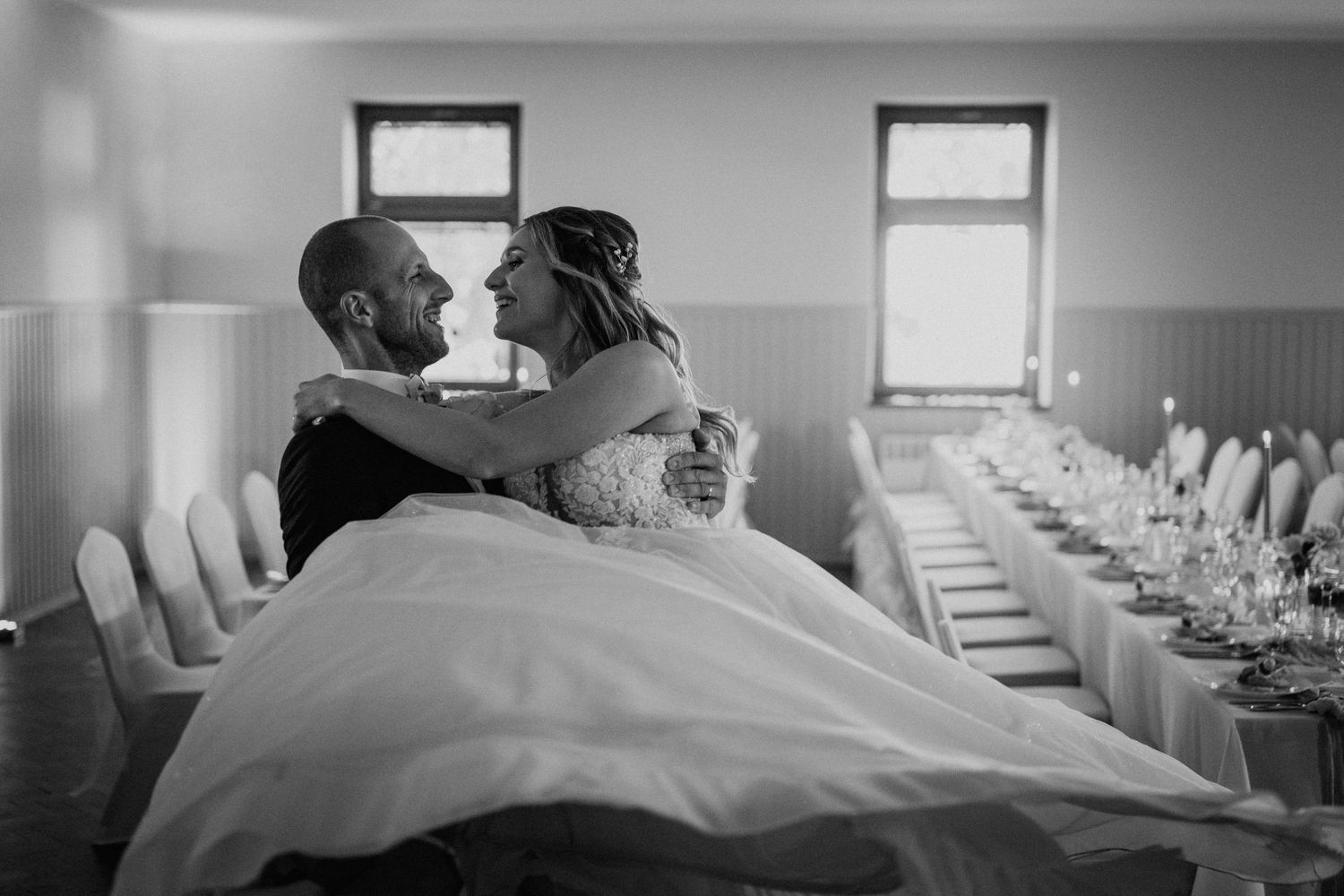 Black and white photo of groom carrying bride in ballgown through decorated wedding reception hall.