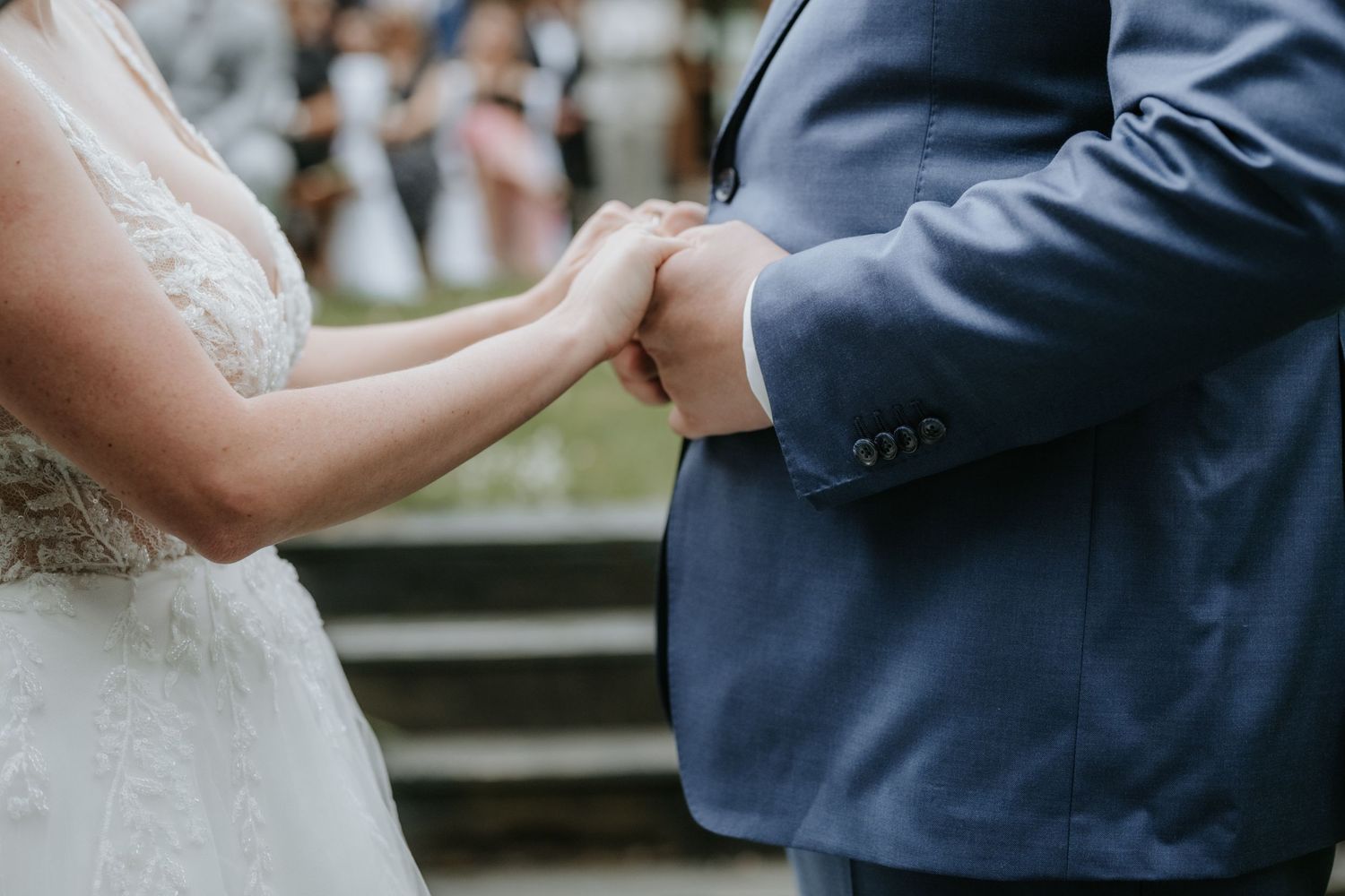 Bride and groom holding hands during wedding ceremony, bride in lace dress and groom in blue suit.