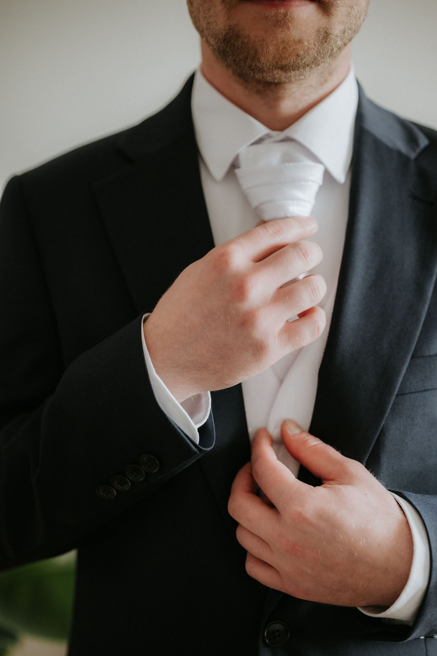Groom in dark suit adjusting white tie while buttoning jacket before wedding.