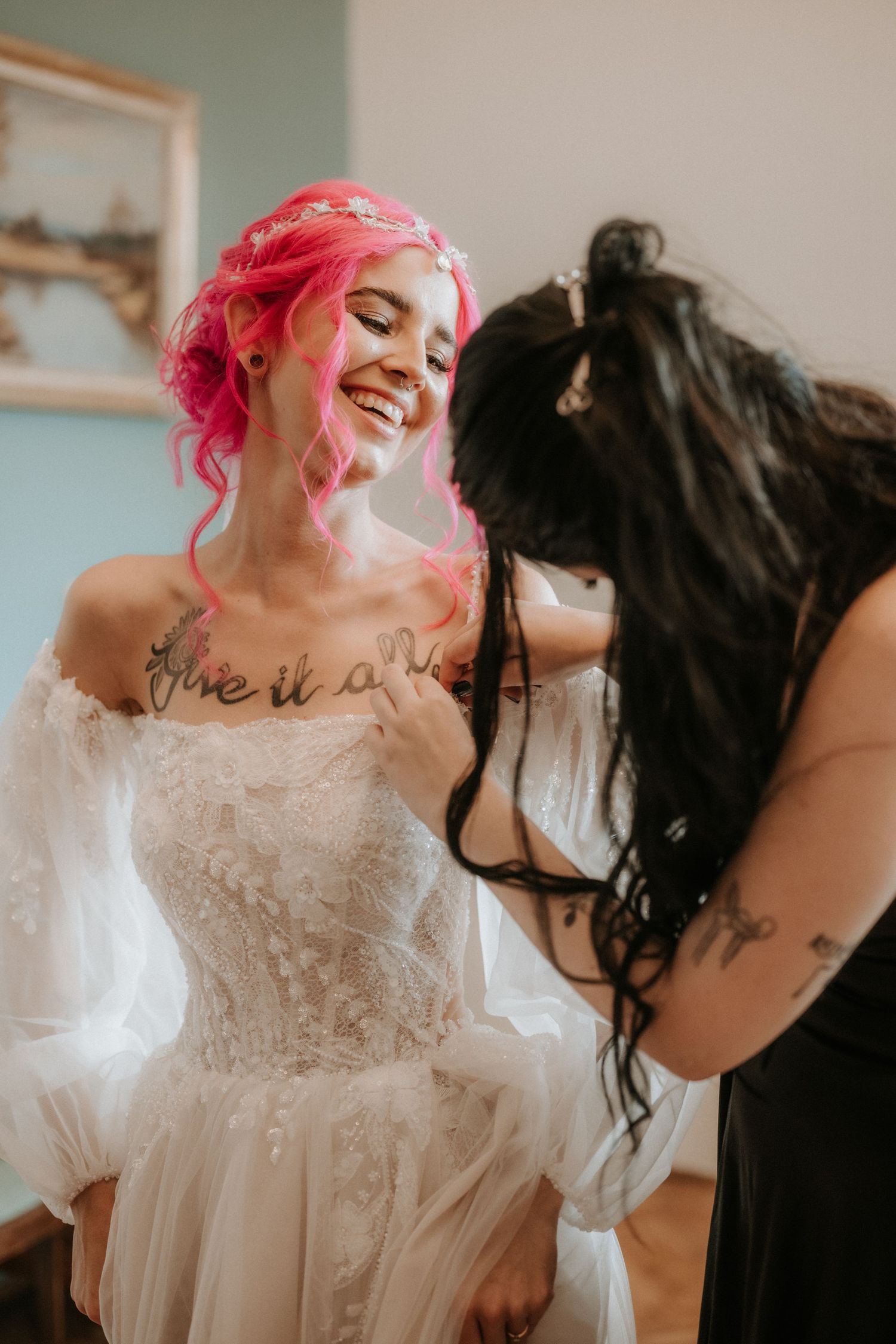 Pink-haired bride smiling in lace wedding gown while bridesmaid with tattoos helps her dress.