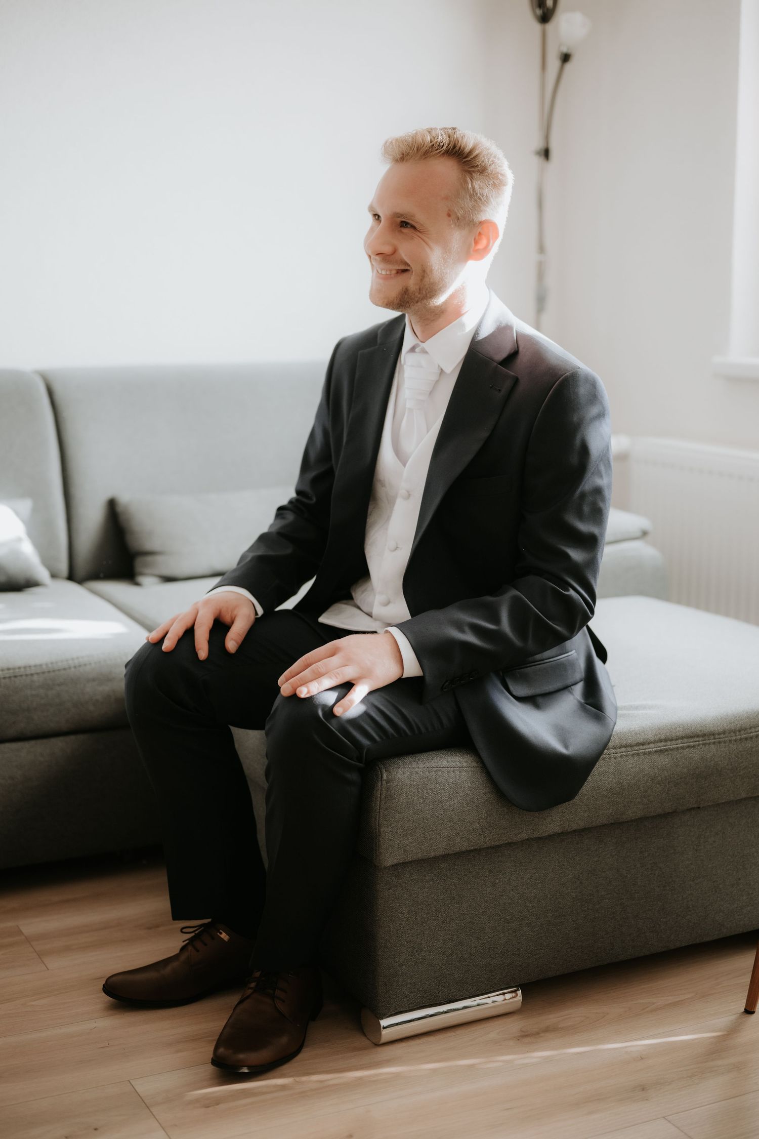 A smiling groom in a black tuxedo sits on a gray sofa, looking away, wearing brown dress shoes on a wooden floor.