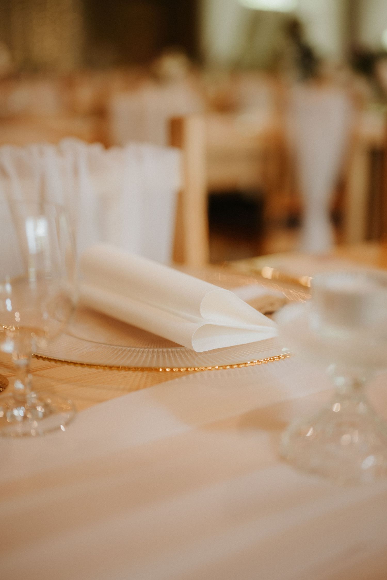 Elegant wedding table setting with white folded napkin on gold beaded charger plate, crystal glassware, and soft bokeh background.