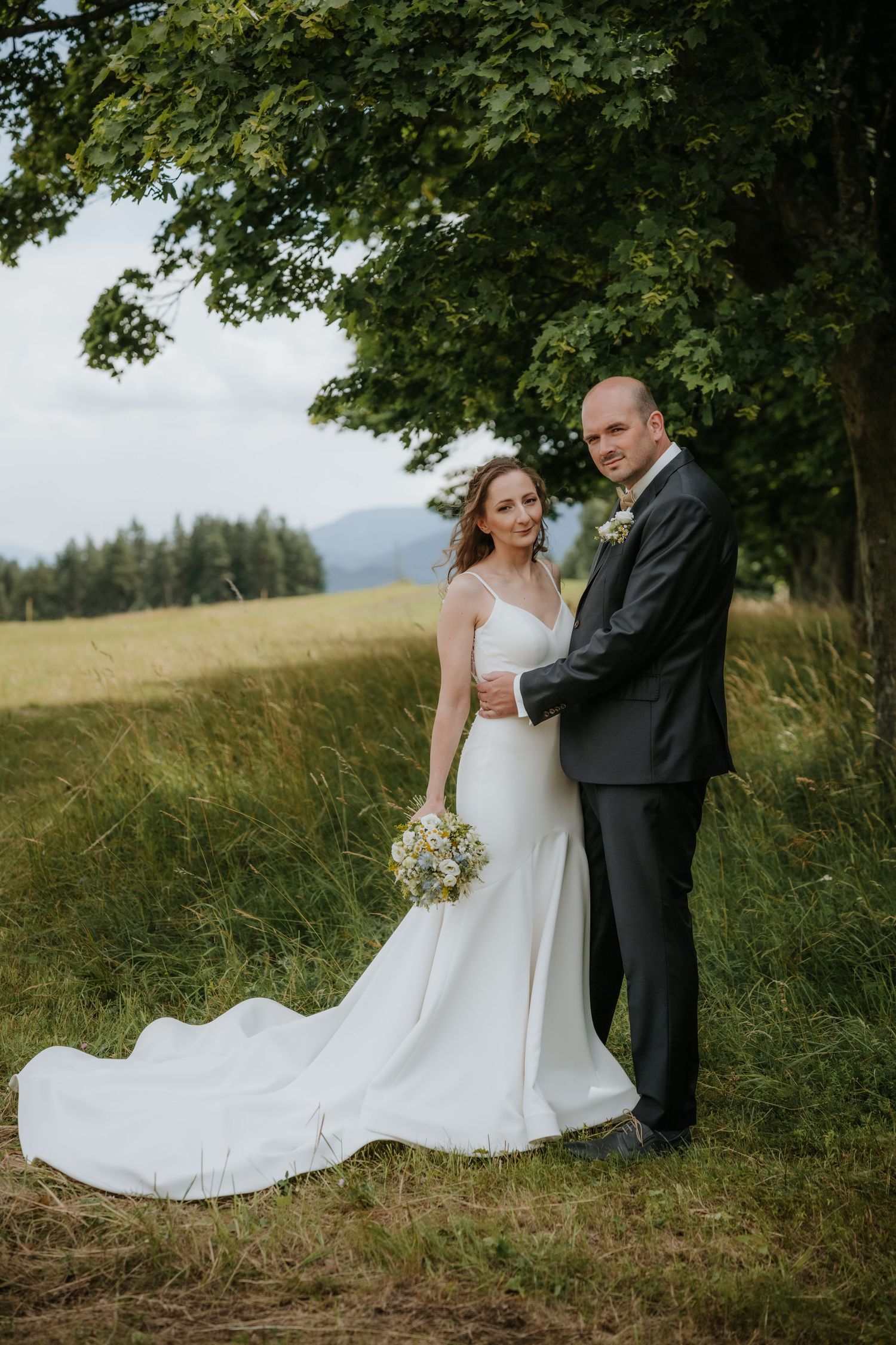 Bride in white gown and groom in gray suit embrace under a large tree in a scenic green meadow.