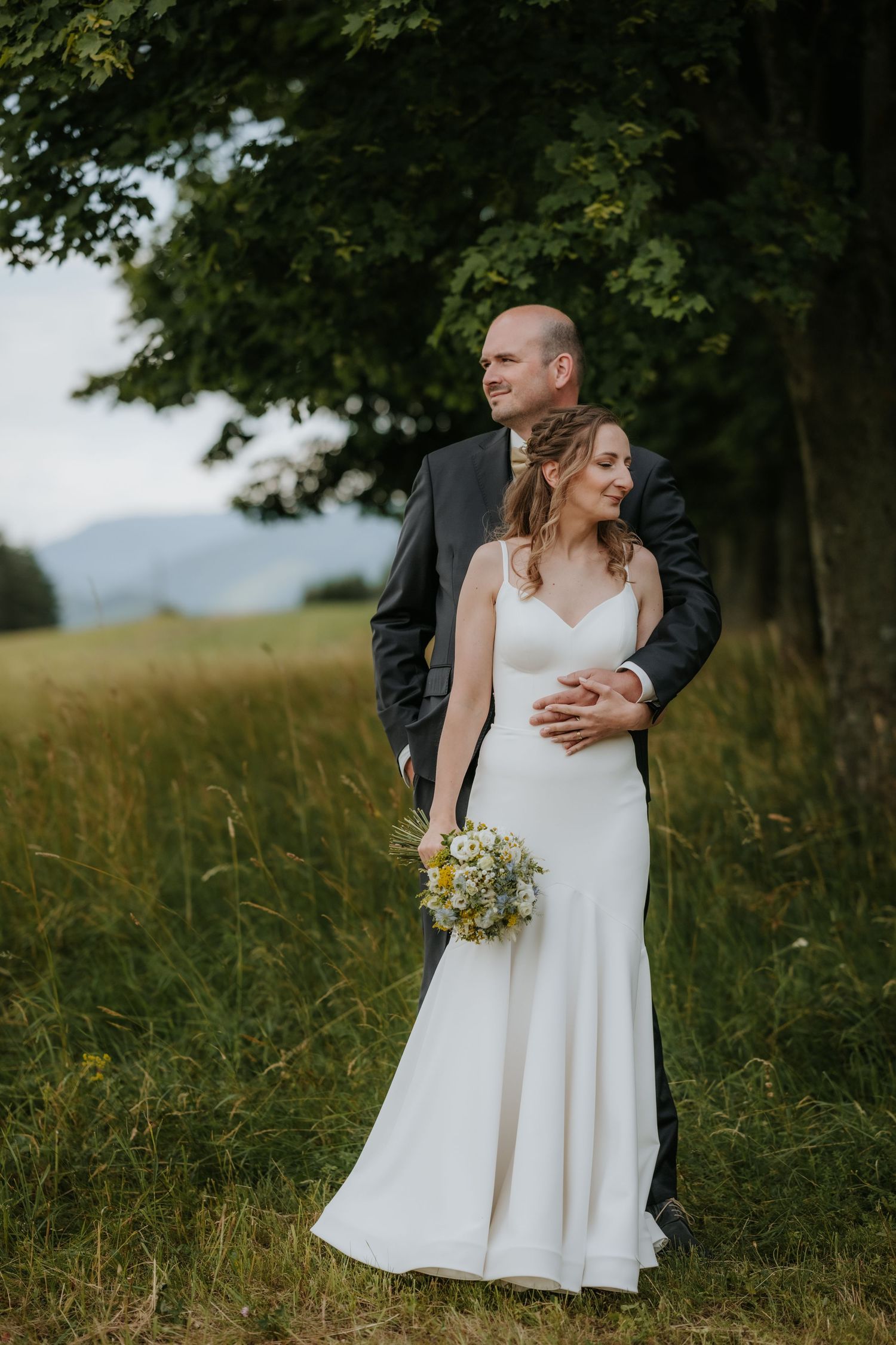 Bride in white gown holding wildflower bouquet embraced by groom in dark suit outdoors near trees and green meadow.