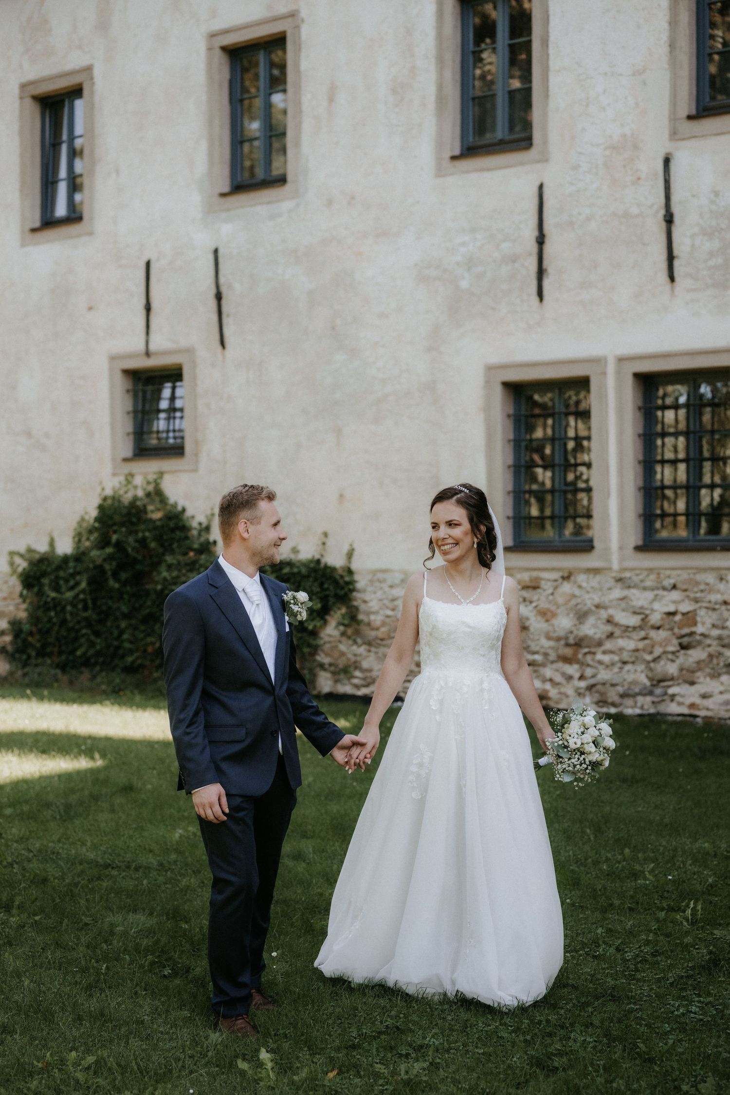 Bride and groom holding hands and walking on green grass outside a historic building.