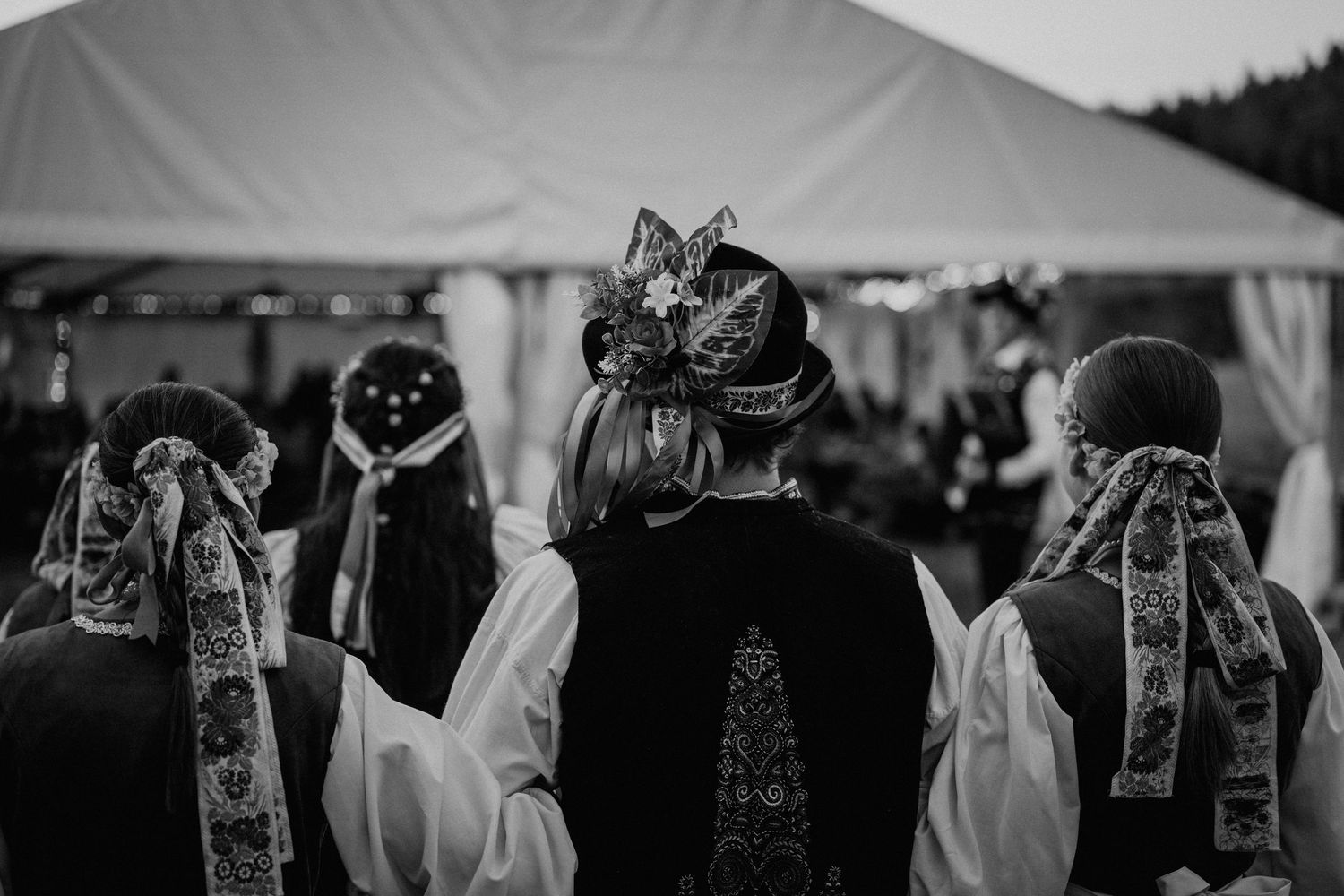 Black and white photo of people in traditional folk costumes with ornate headdresses at an outdoor event.