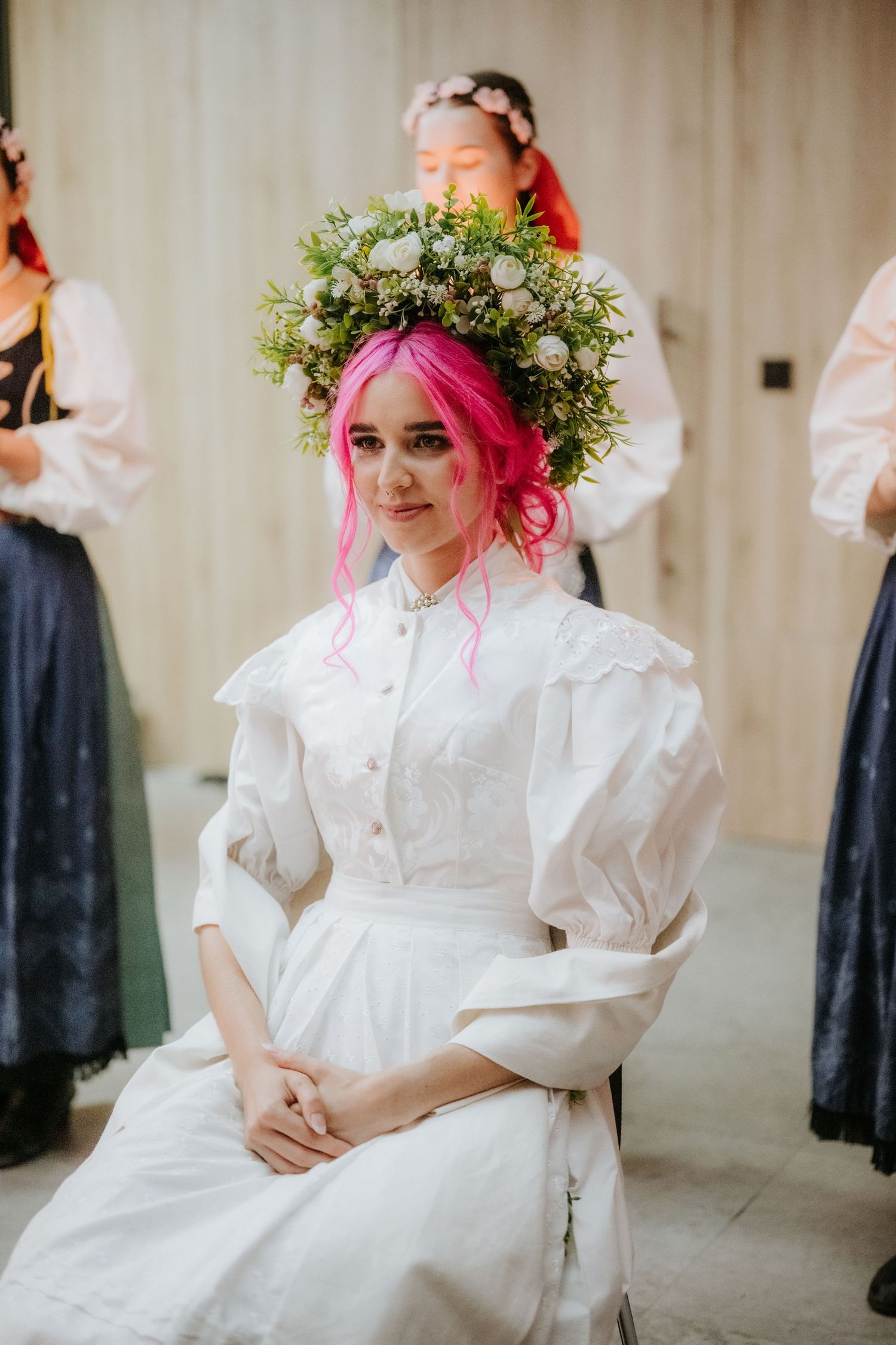 Woman with pink hair wearing a white dress and floral crown, seated while another person arranges her hair.