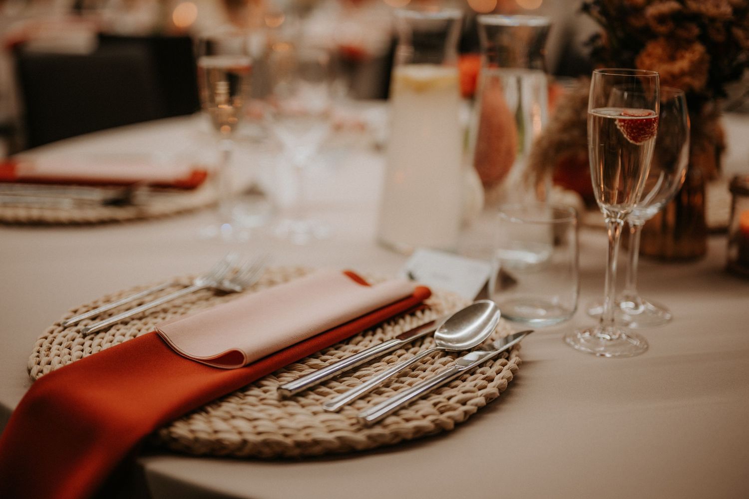 Elegant wedding table setting with woven charger plate, silverware, terracotta napkin, and crystal wine glasses.