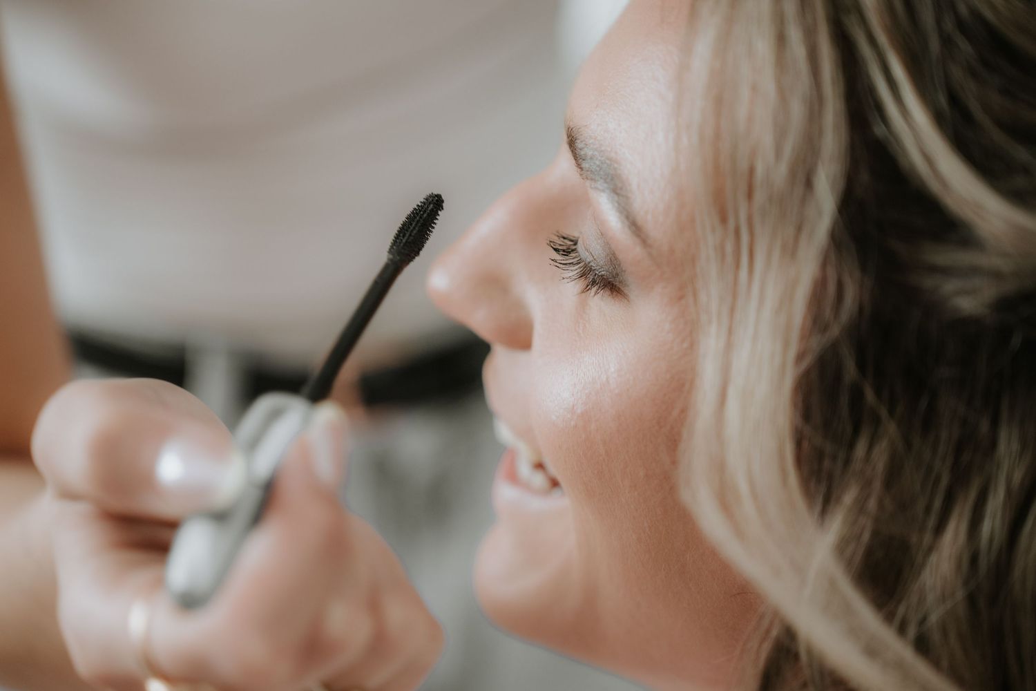 Close-up of a smiling woman applying mascara with a wand, showcasing a natural glam makeup look.