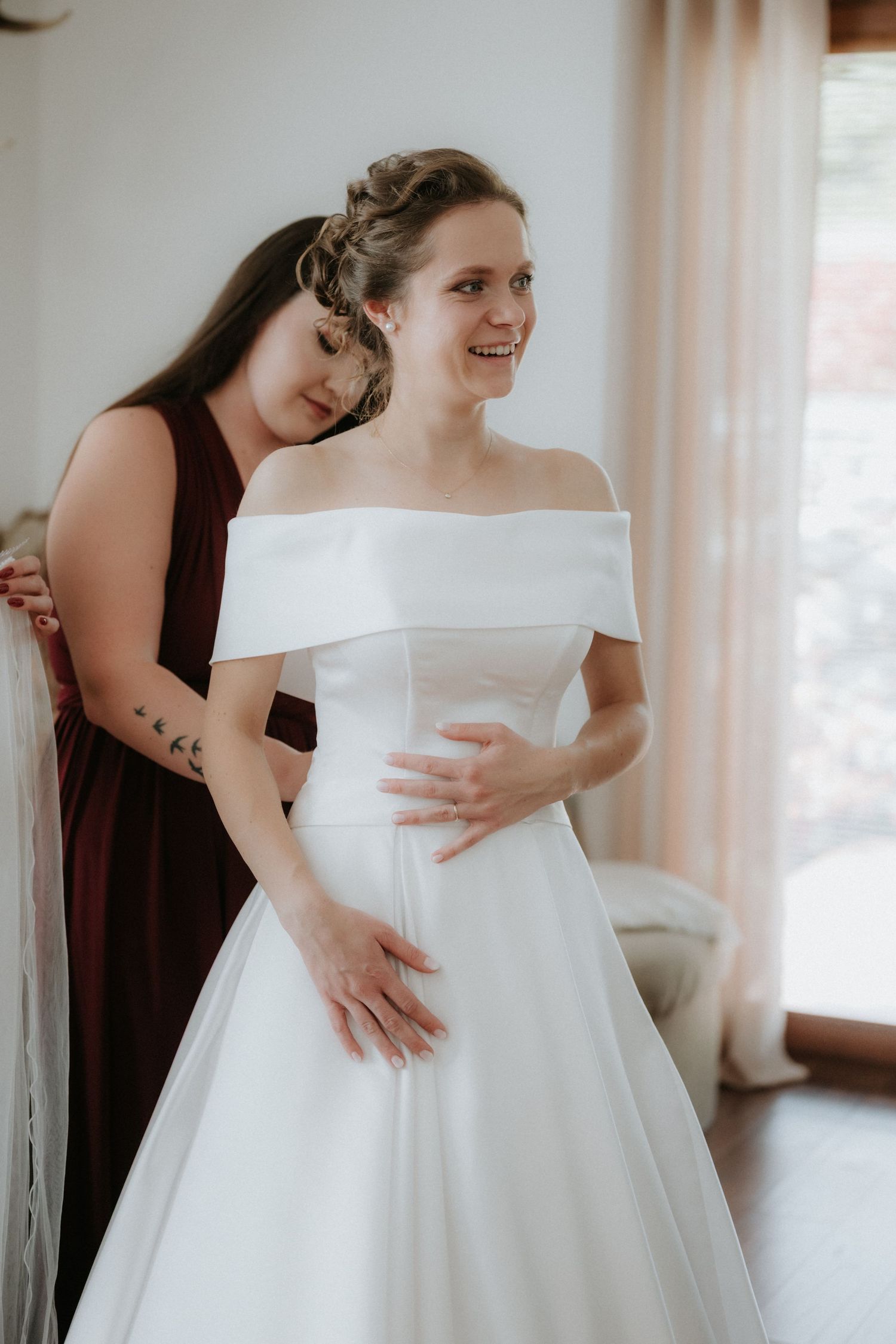 Bridesmaid in burgundy dress helping bride into elegant off-shoulder white wedding gown in bright room.