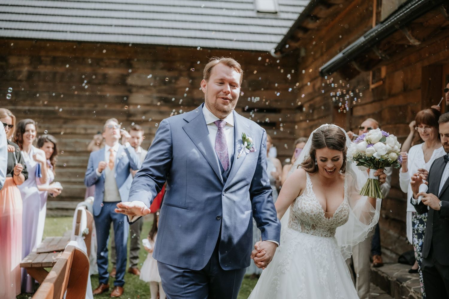 Bride and groom walk hand in hand down the aisle as guests celebrate with bubbles at outdoor wedding.