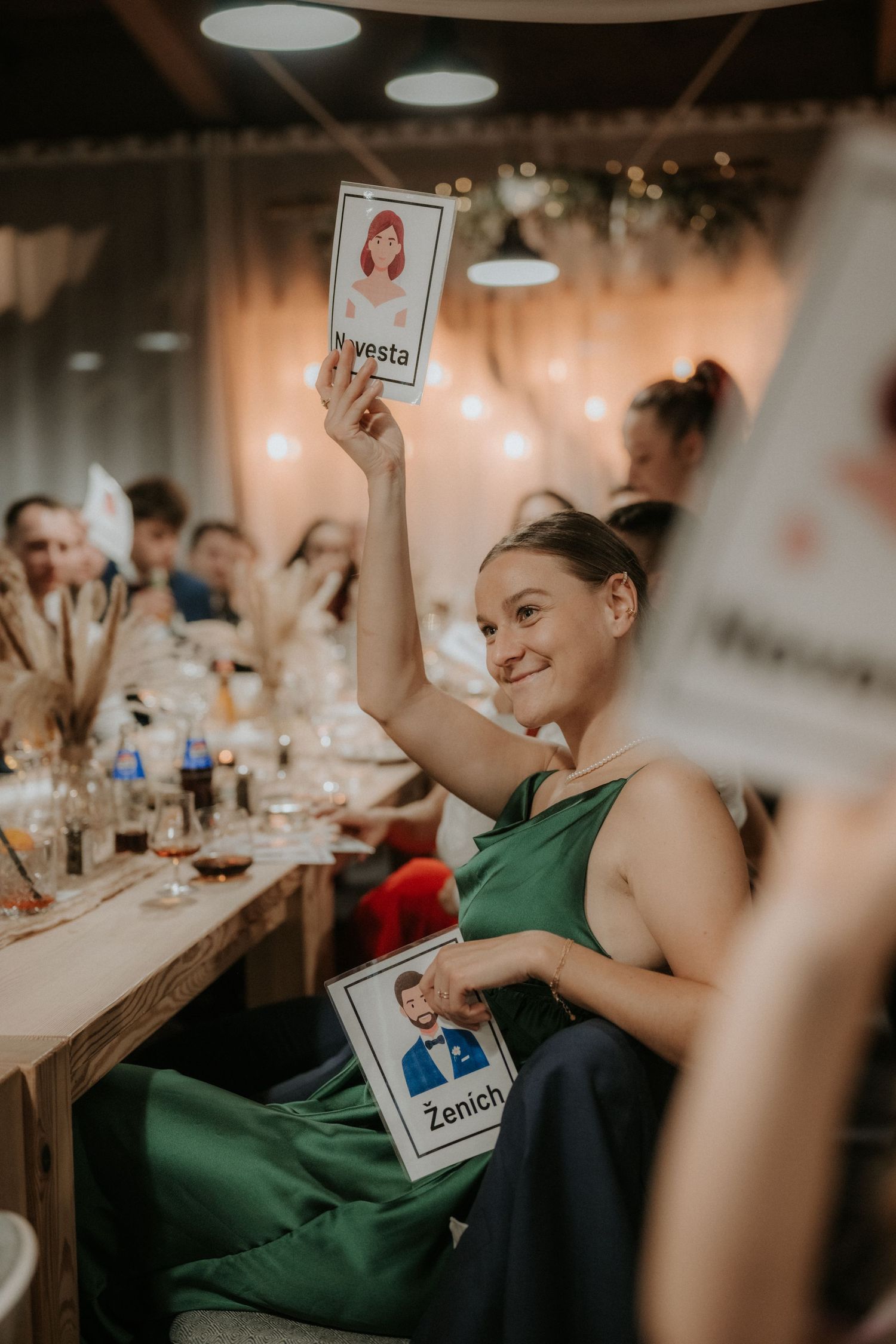 Woman in green dress holds up Nevesta and Zenich cards at a wedding reception game.