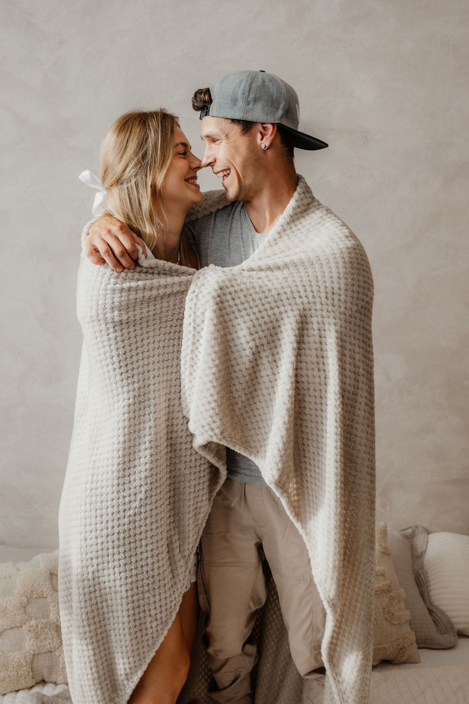 Happy couple wrapped in a cozy white waffle blanket, smiling at each other on a bed.