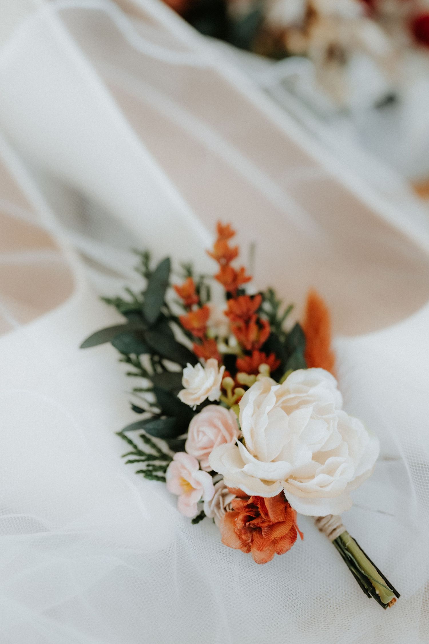 Elegant wedding boutonniere with white peony, orange flowers, and greenery resting on a bridal veil.