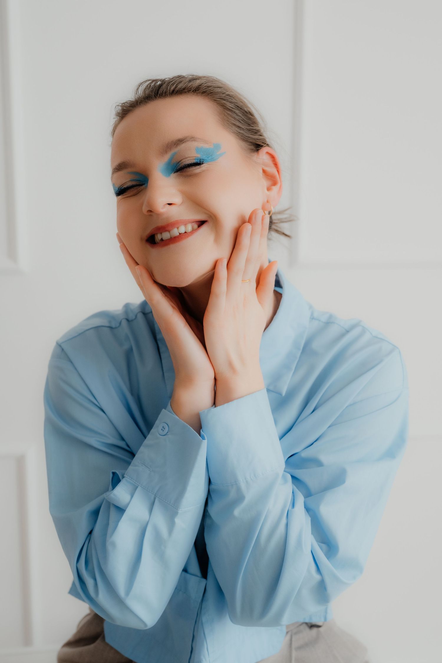 Smiling woman with blue eye makeup in blue shirt touching her face with both hands.
