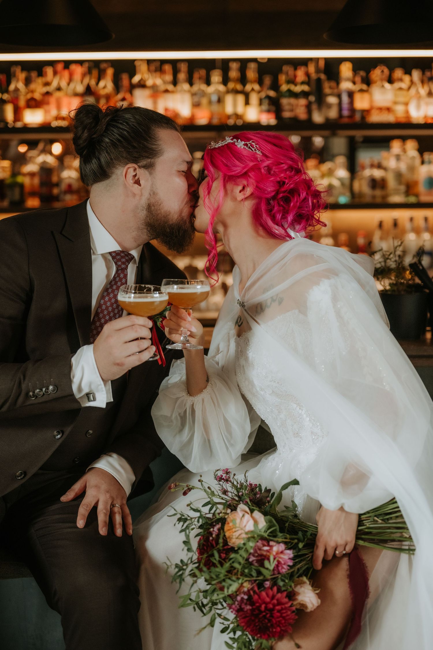 Pink-haired bride and groom sharing a kiss while holding drinks at a bar with floral bouquet.