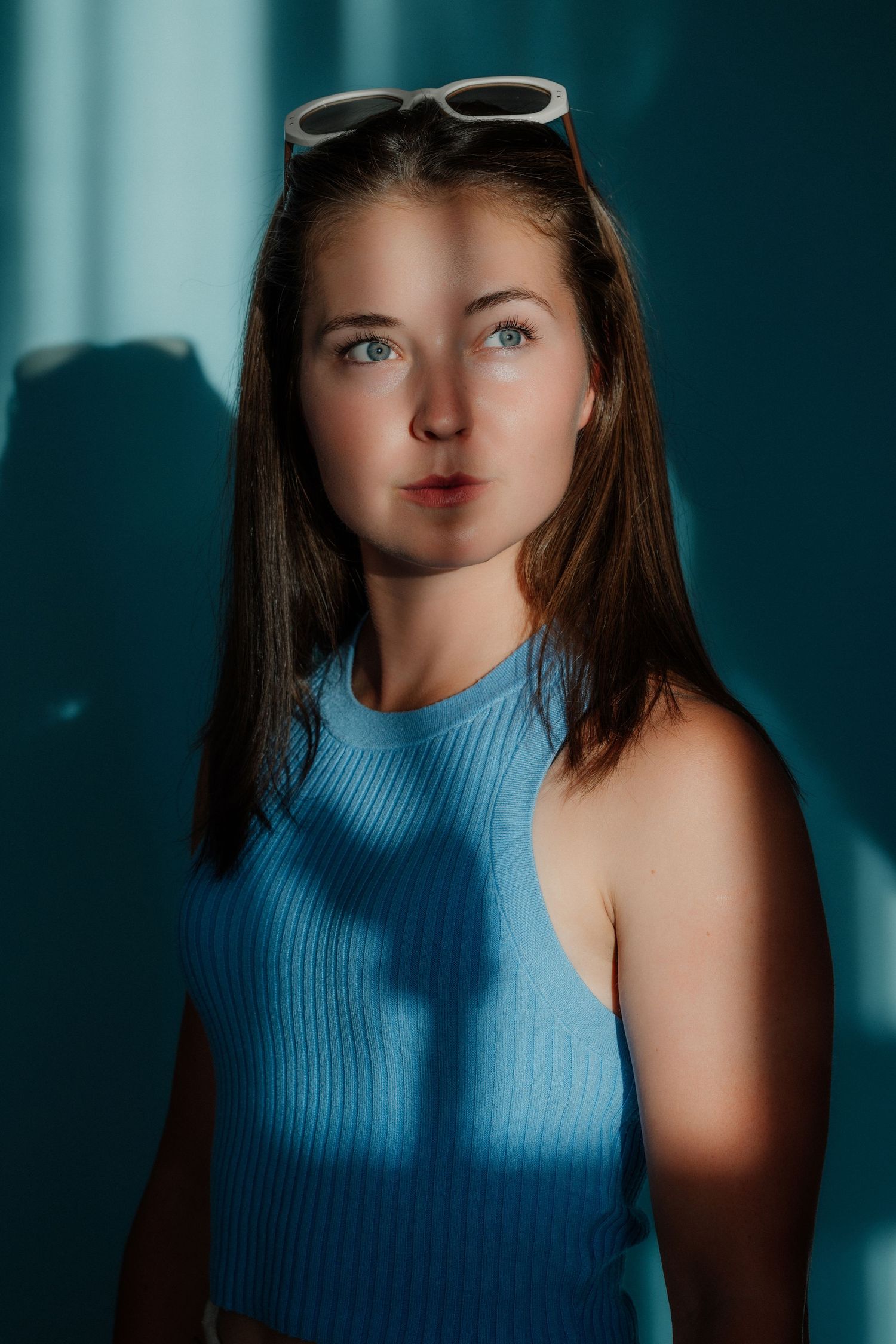 Young woman with blue eyes wearing a blue ribbed tank top and white sunglasses on her head in dramatic lighting.