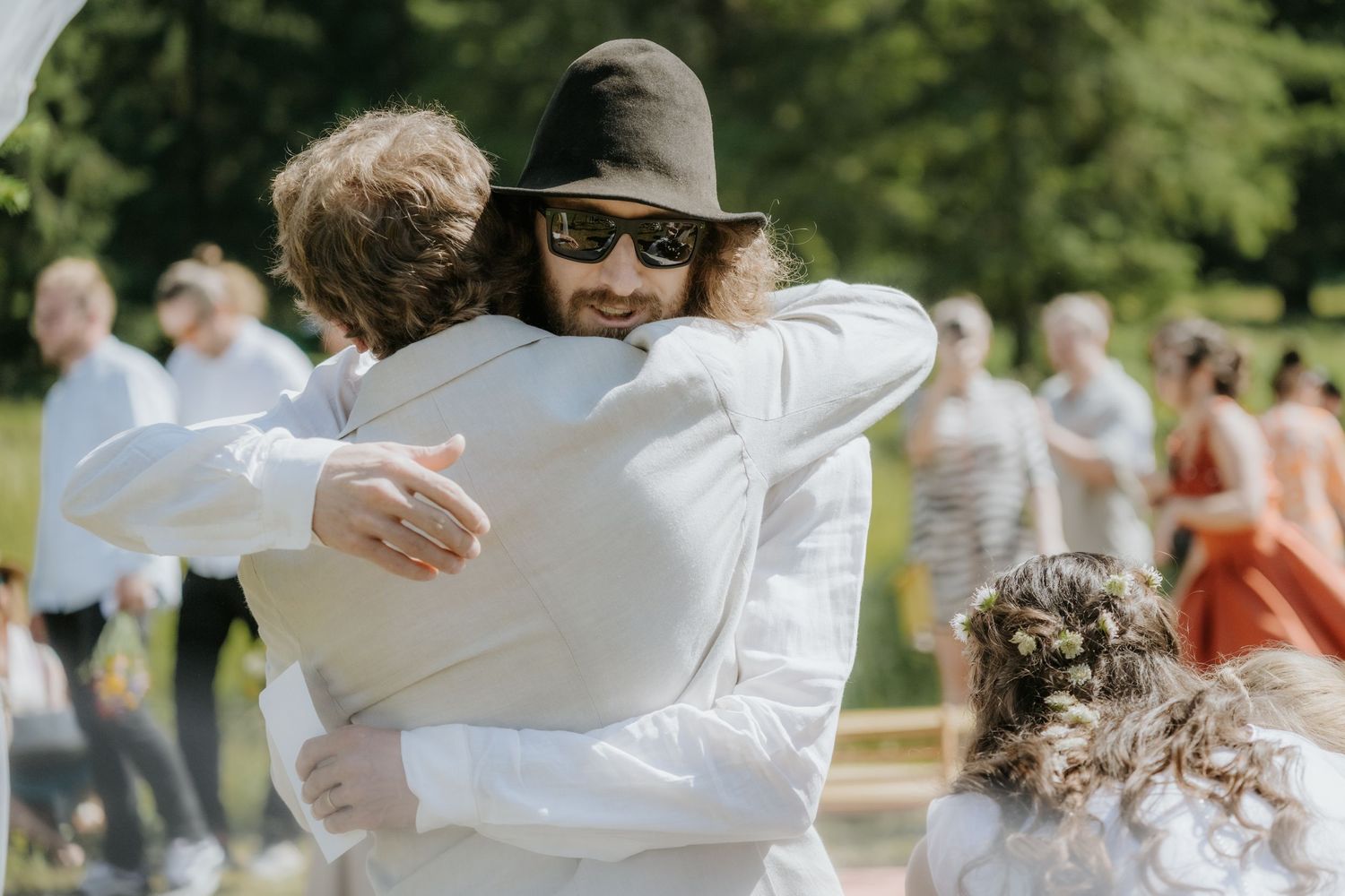 Two men in white shirts share a warm hug at an outdoor wedding ceremony on a sunny day.