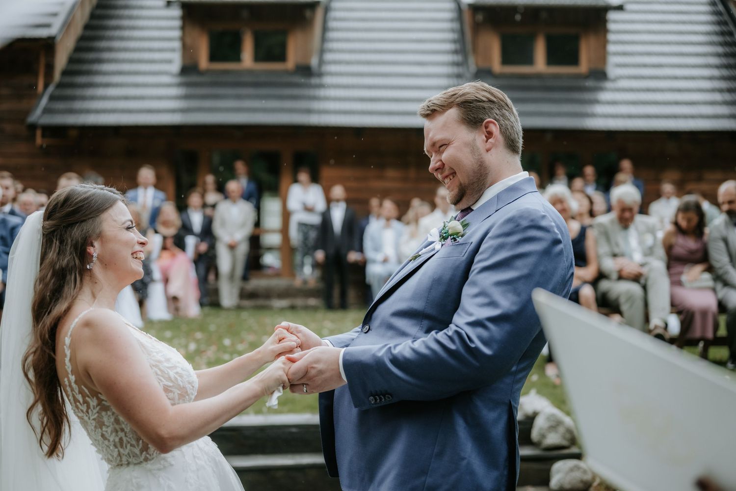Groom places wedding ring on bride's finger during outdoor ceremony with guests seated behind rustic wooden venue.