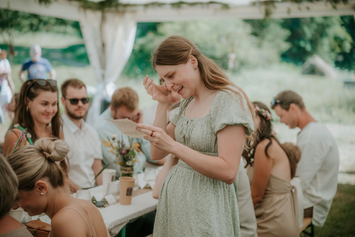 A woman in a green floral dress laughs joyfully at an outdoor wedding reception under a white tent.
